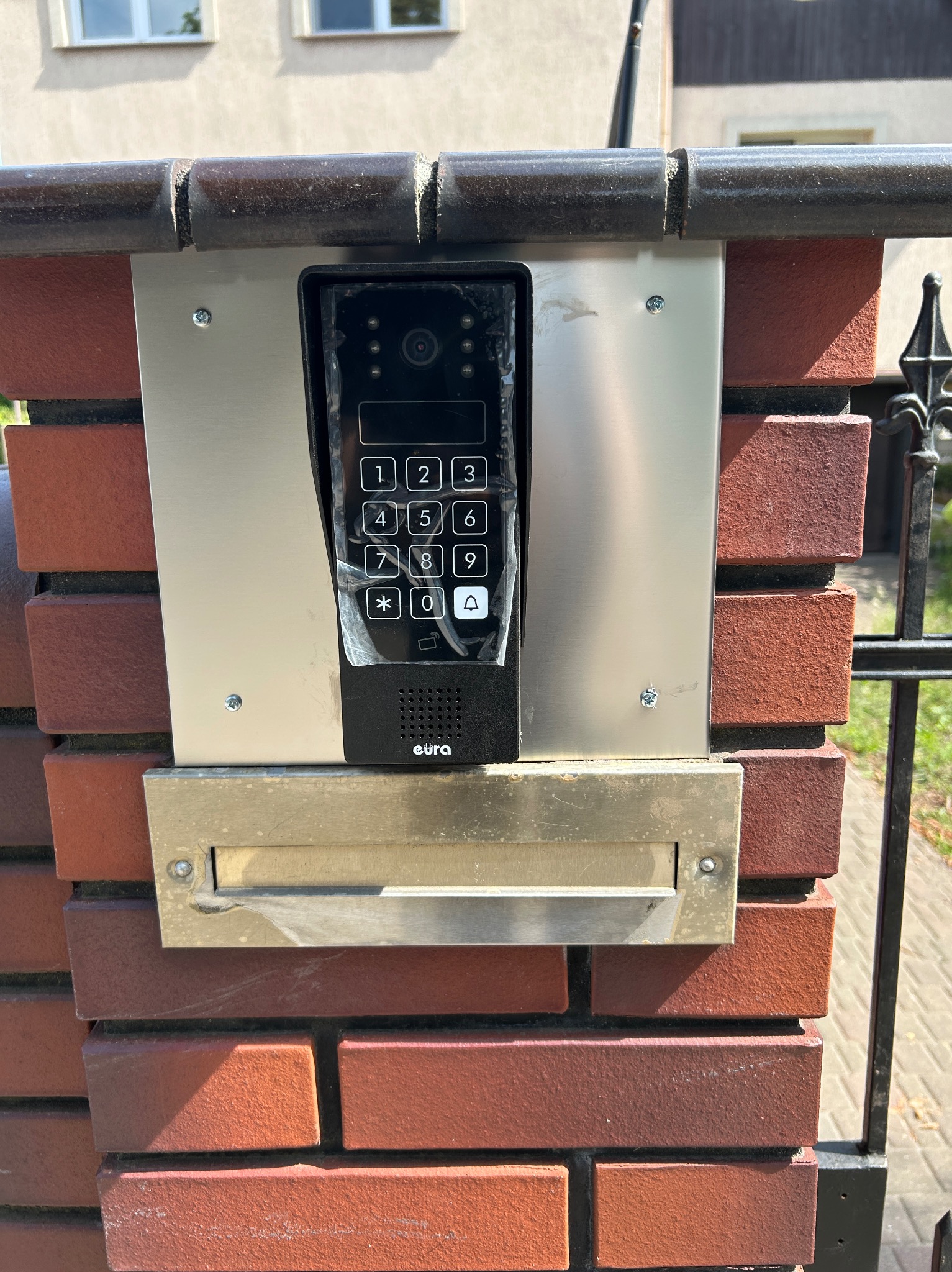 Zamontowany wideodomofon EURA z klawiaturą numeryczną, umieszczony na ceglanym słupku ogrodzenia z skrzynką na listy. Widoczne śruby mocujące i fragment metalowej bramy.