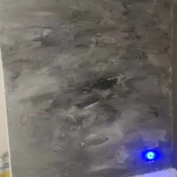 Ściana pokryta szarą, dekoracyjną gładzią z błyszczącymi drobinkami, sąsiadująca z białym kominkiem, oświetlona niebieskim światłem z boku.