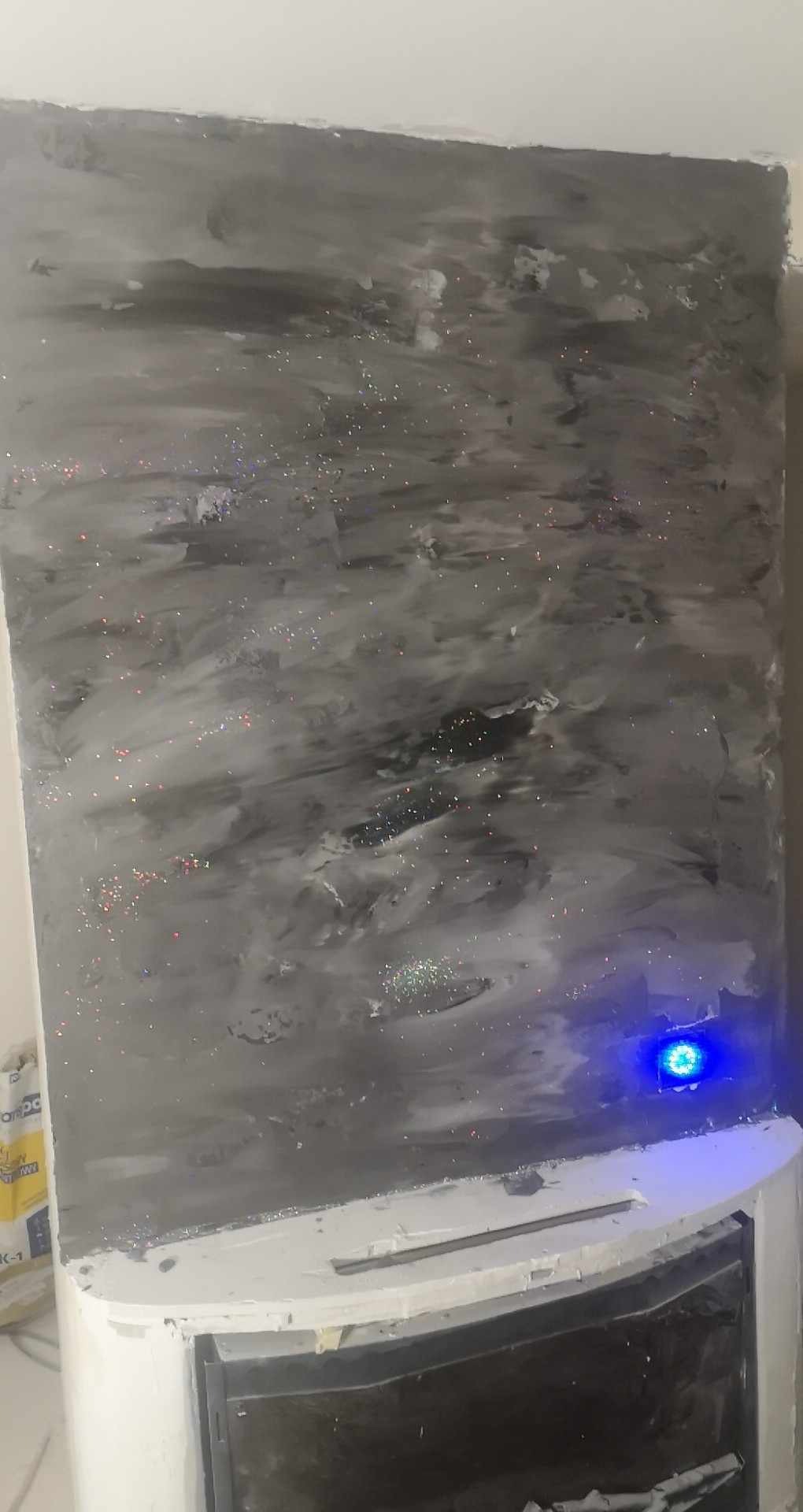 Ściana pokryta szarą, dekoracyjną gładzią z błyszczącymi drobinkami, sąsiadująca z białym kominkiem, oświetlona niebieskim światłem z boku.