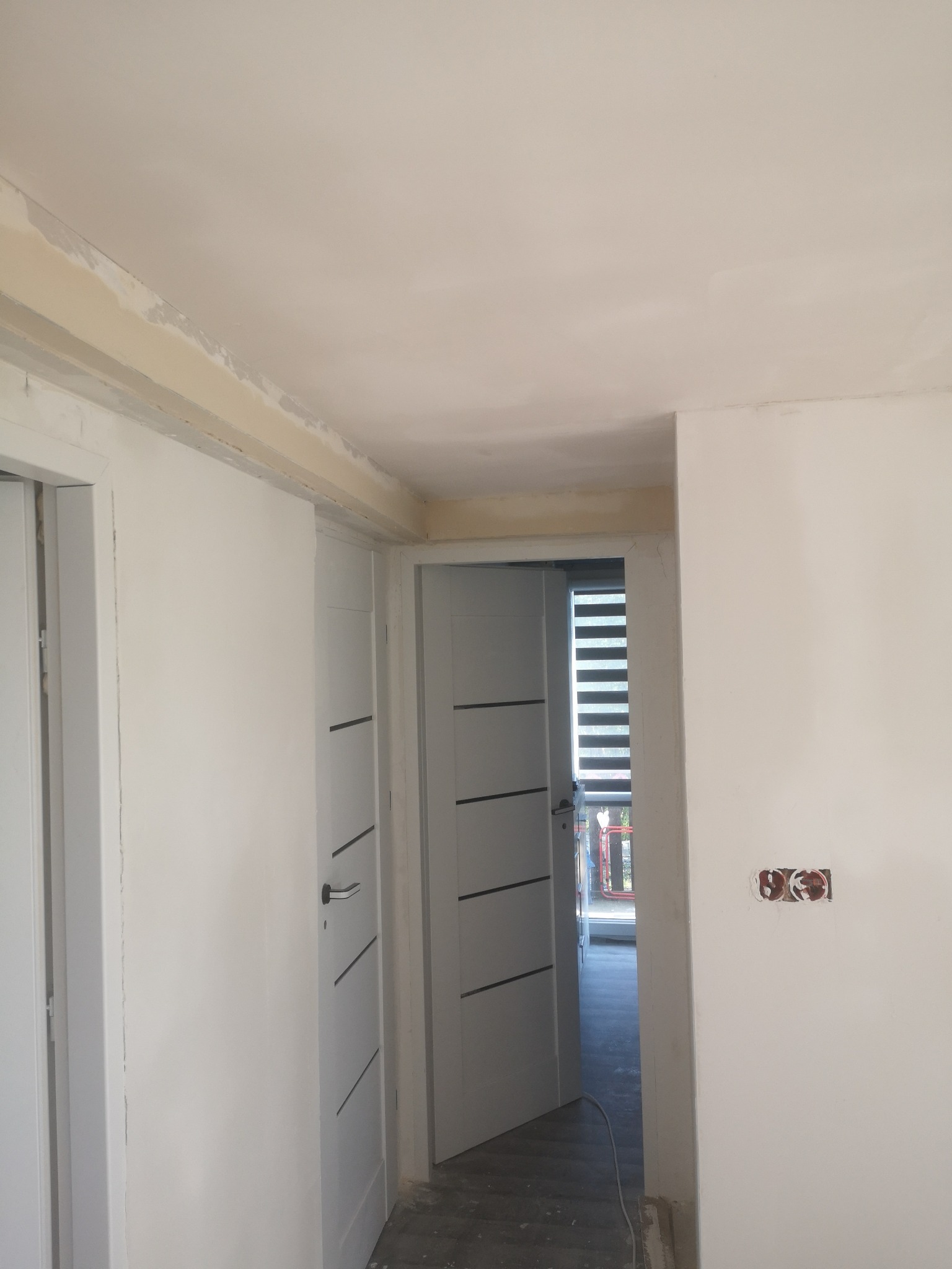 Wnętrze w trakcie prac: częściowo wyszpachlowany sufit z widocznymi łączeniami, białe ściany, otwarte białe drzwi z czarnymi akcentami i wystające kable ze ściany.