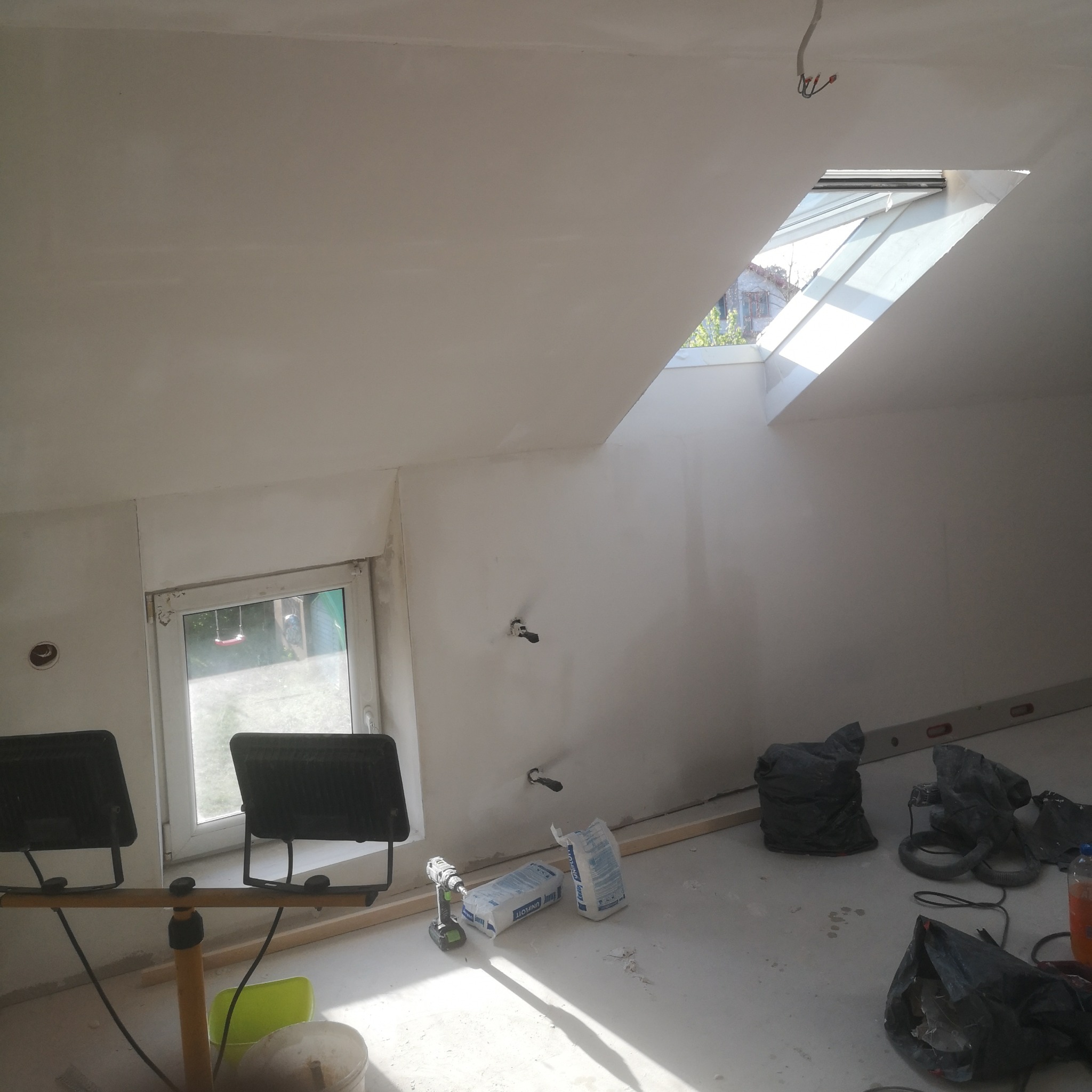 Poddasze w trakcie prac wykończeniowych: świeżo położona biała warstwa na ścianach i suficie, okno dachowe wpuszczające jasne światło, narzędzia i materiały budowlane rozłożone na podłodze, w tym...