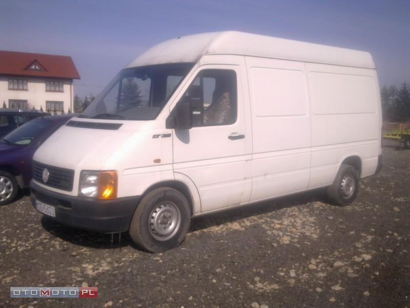 Biały samochód dostawczy Volkswagen LT 35 na żwirowym parkingu, widoczny z boku, z fragmentem innego auta i budynku w tle.