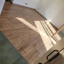 Ułożone panele podłogowe w nowym pomieszczeniu, widoczne promienie słoneczne, niebieska taśma malarska przy ścianach oraz fragment szafki w rogu kadru.