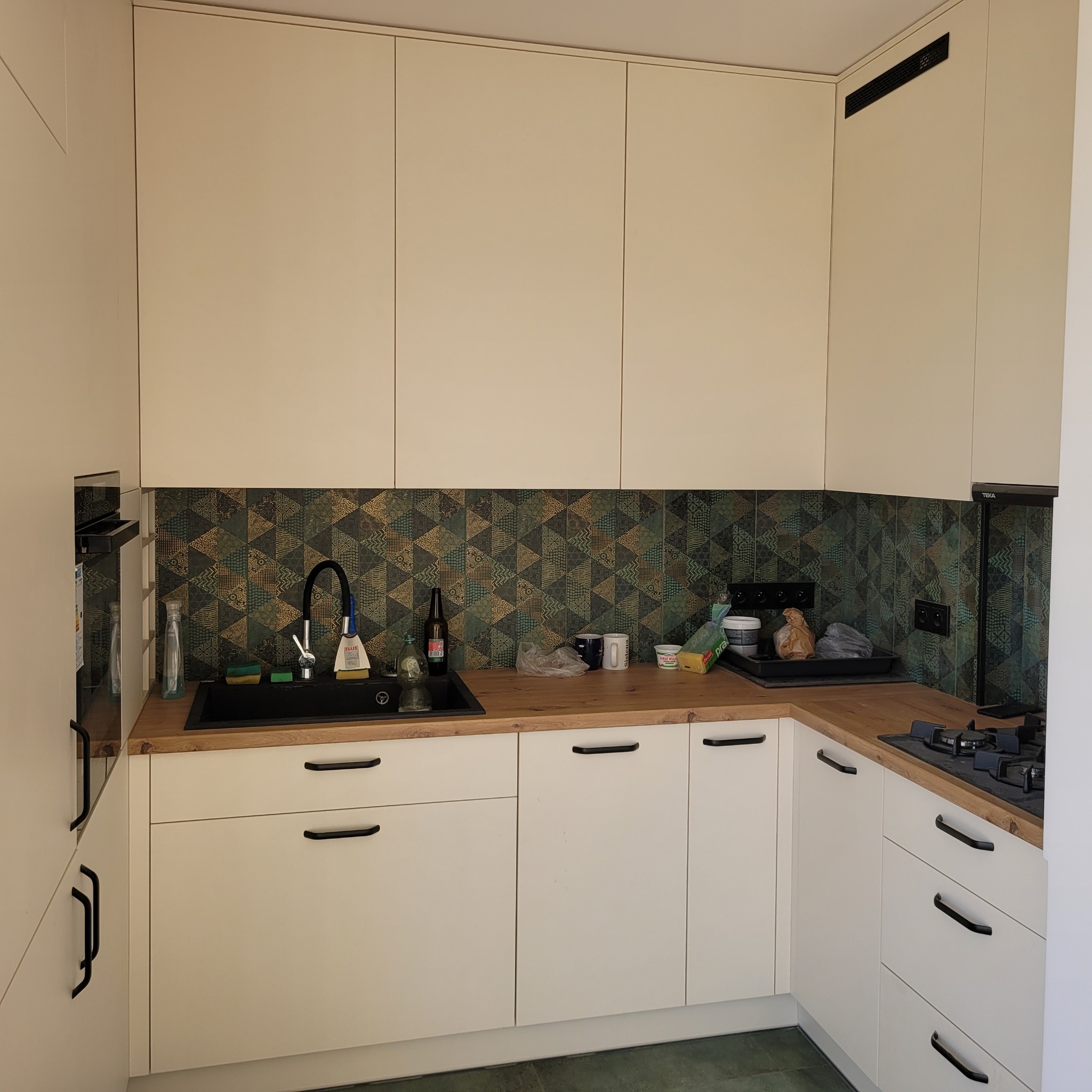 Nowoczesna kuchnia z białymi szafkami, drewnianym blatem i czarnym zlewem oraz geometrycznymi płytkami na ścianie.