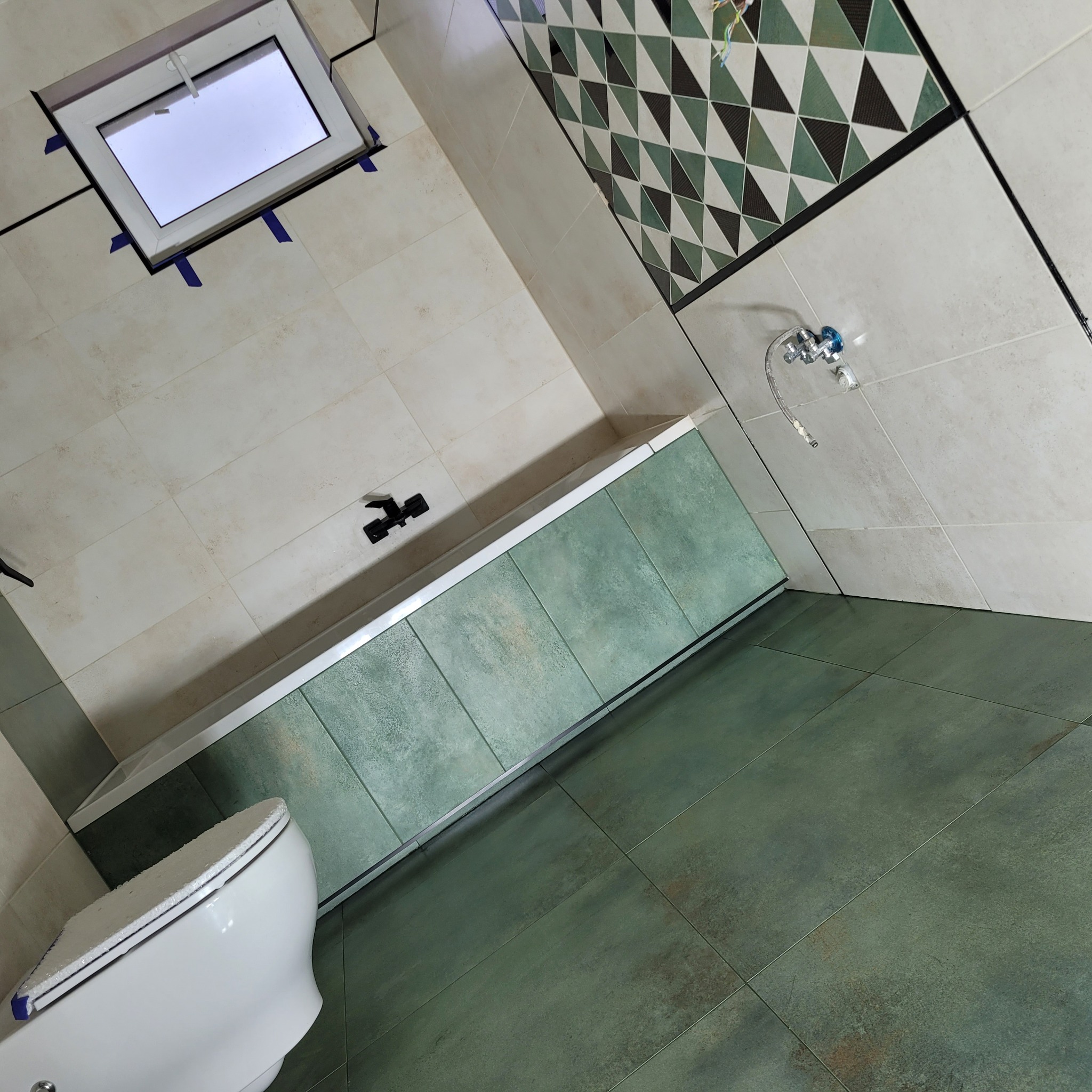 Nowoczesna łazienka w Wieliszewie po remoncie: zielono-beżowe płytki, geometryczny wzór nad wanną, biała armatura, okno dachowe zabezpieczone taśmą.