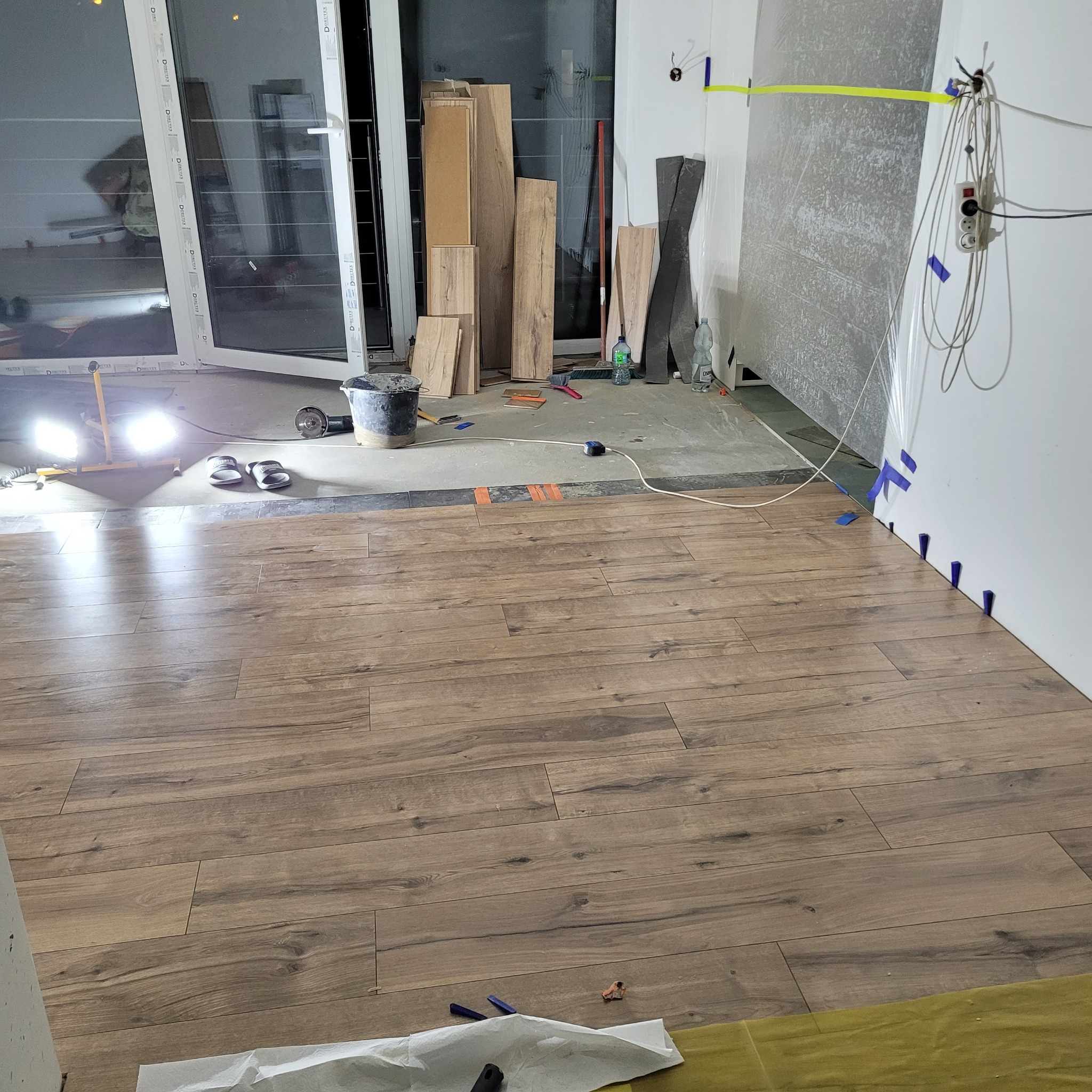 Układanie paneli podłogowych imitujących drewno w trakcie prac wykończeniowych, widoczne kliny dystansowe przy ścianie, narzędzia i materiały budowlane w tle oraz oświetlenie robocze.