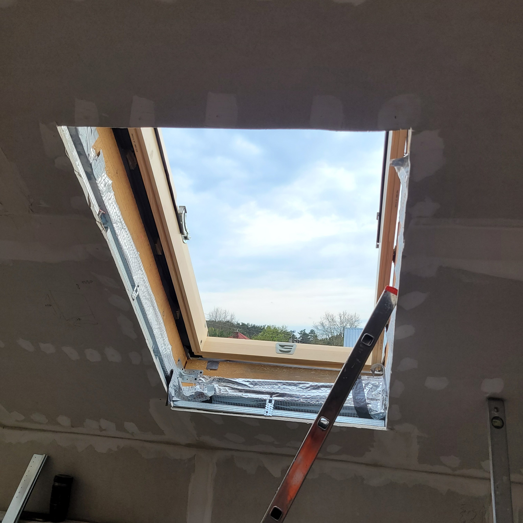 Montaż okna dachowego, widoczne ocieplenie folią aluminiową, częściowo położona gładź, drabina oparta o ścianę, oraz poziomica.