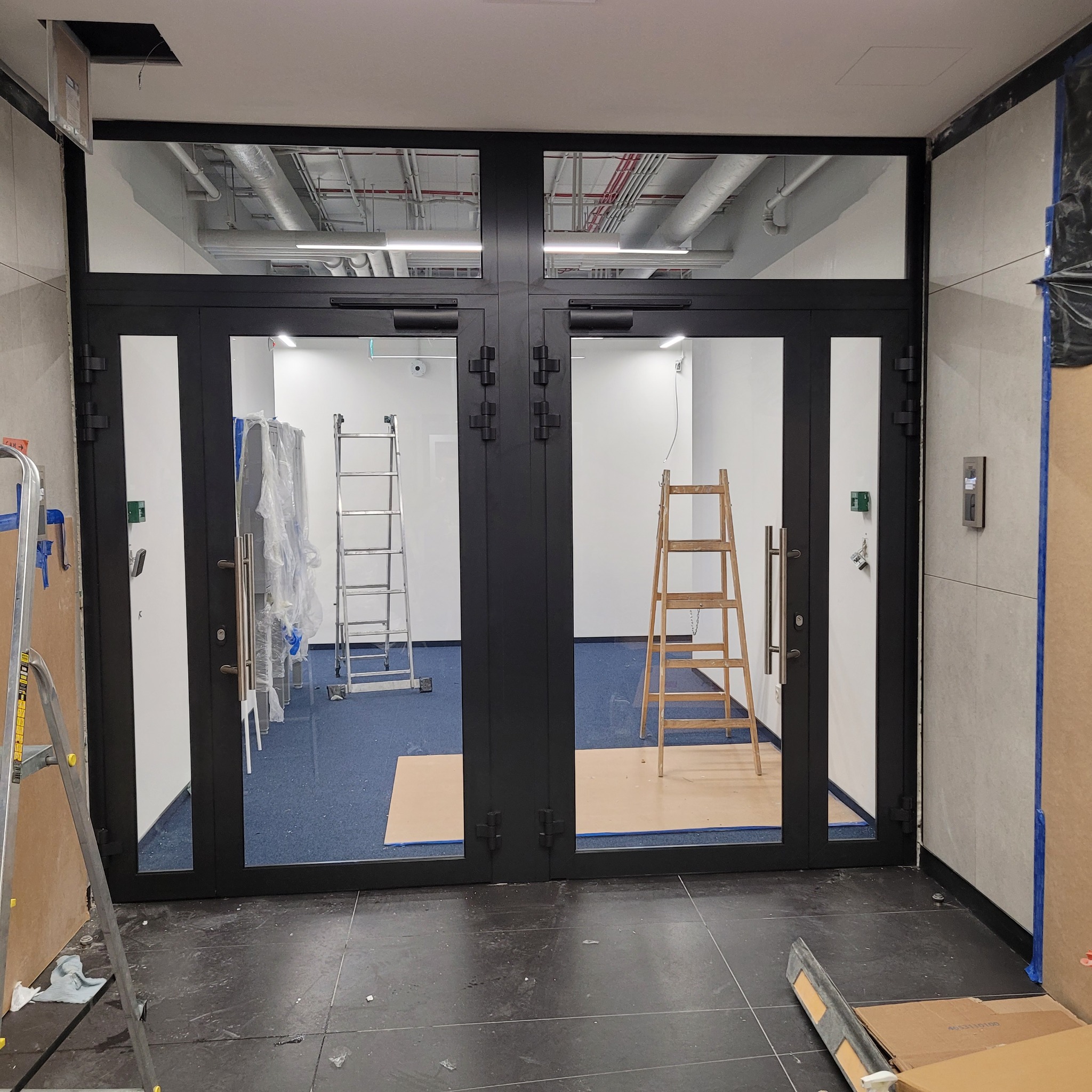 Montaż dwuskrzydłowych, czarnych drzwi z przeszkleniami w lokalu biurowym, widoczne drabiny i zabezpieczenia podłogi.