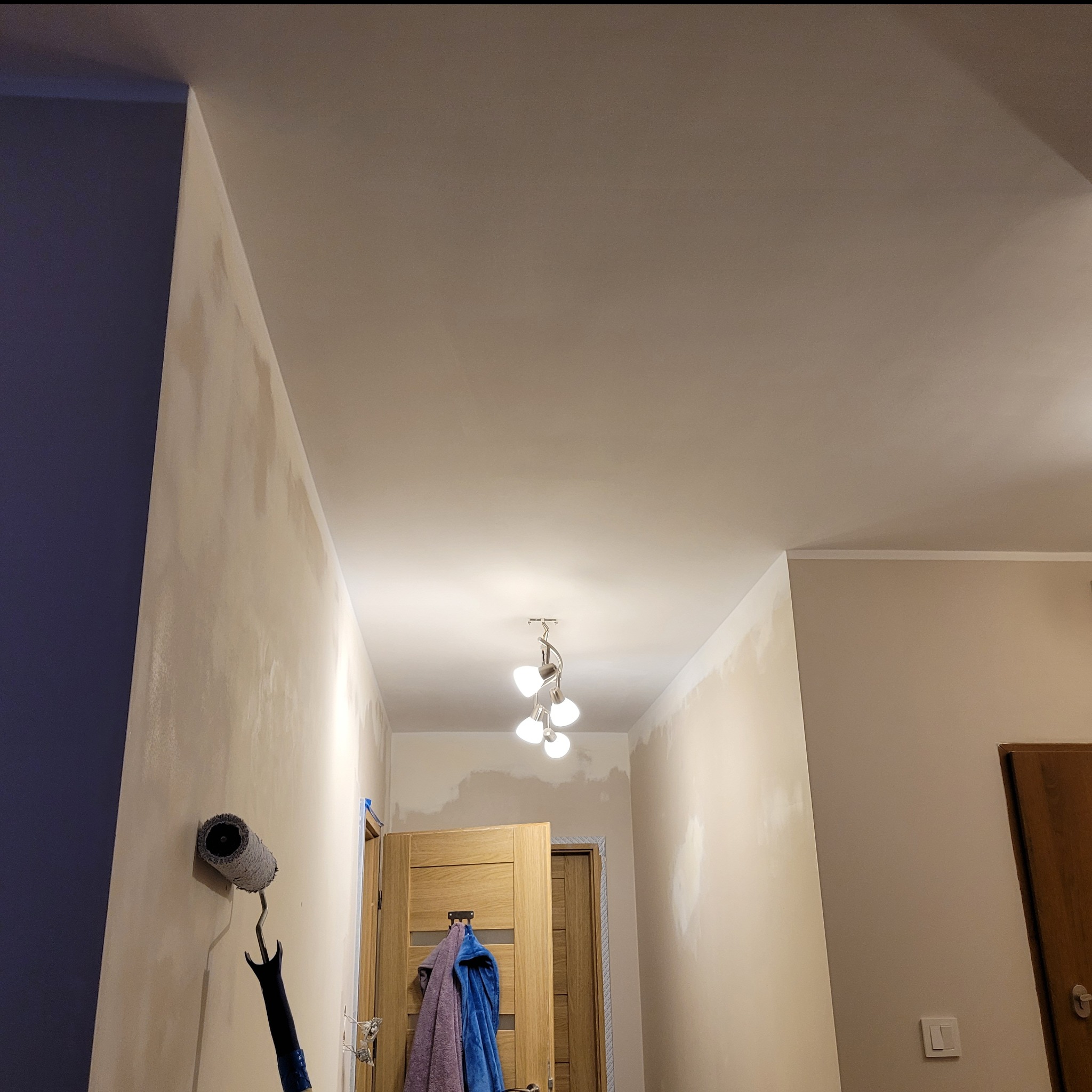 Korytarz w trakcie malowania, ściany z widocznymi plamami po nałożeniu pierwszej warstwy, wałek do malowania oparty o ścianę, widoczne oświetlenie sufitowe.