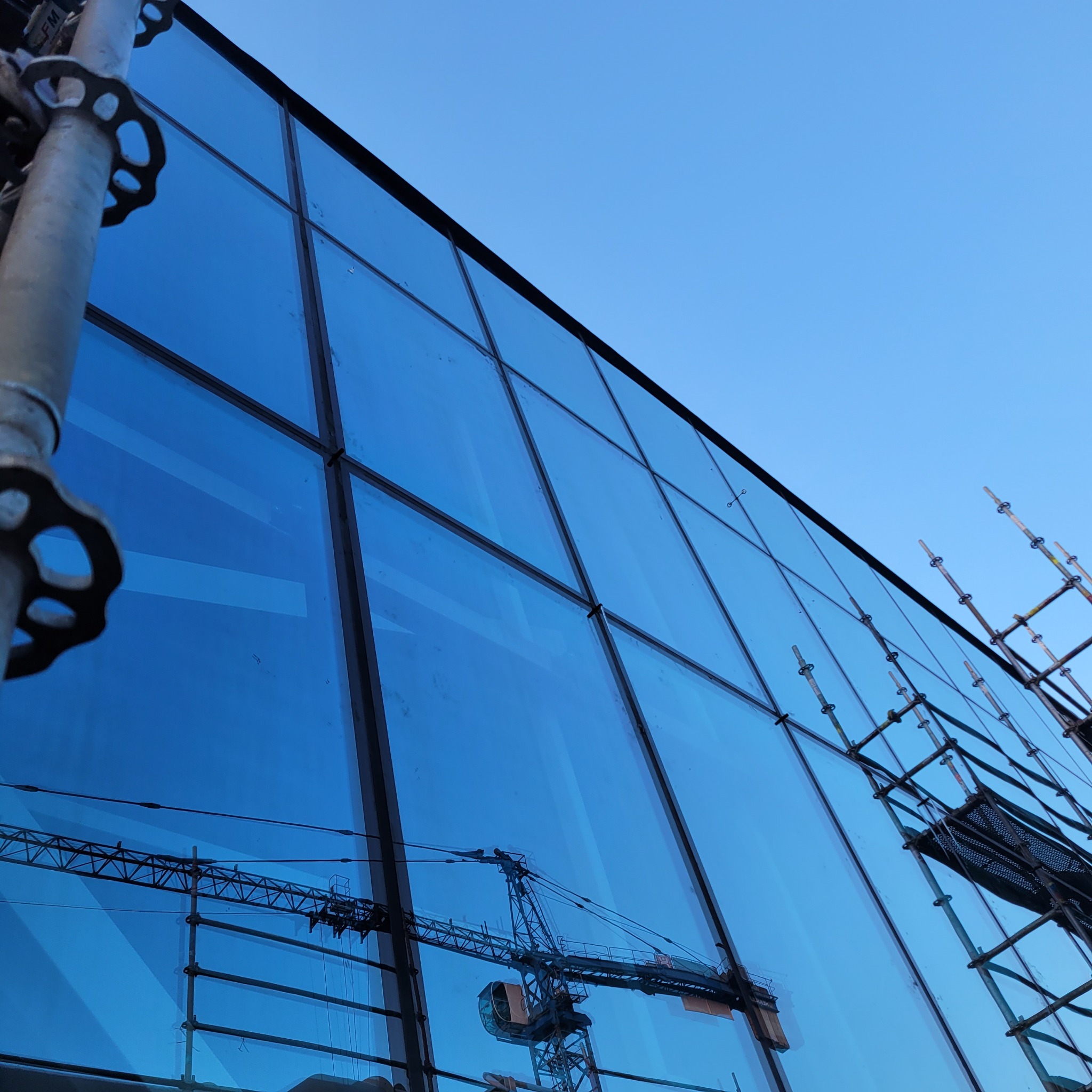 Wysoka szklana fasada budynku w trakcie montażu, widoczne rusztowania i odbicie żurawia budowlanego na powierzchni szyb.