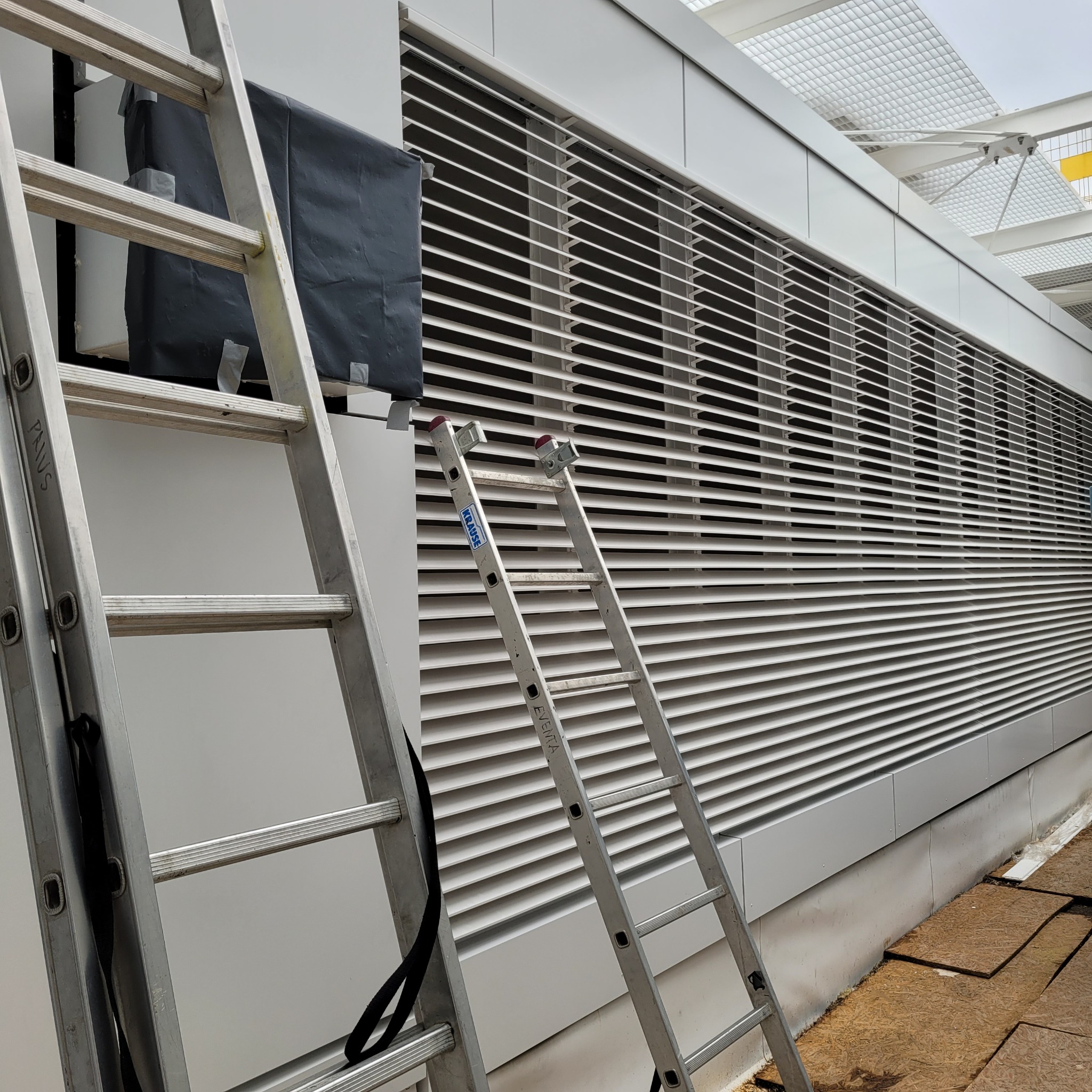 Dwie aluminiowe drabiny stojące przed długą, poziomą żaluzją na elewacji budynku; jedna z sekcji żaluzji zabezpieczona czarną folią i taśmą.