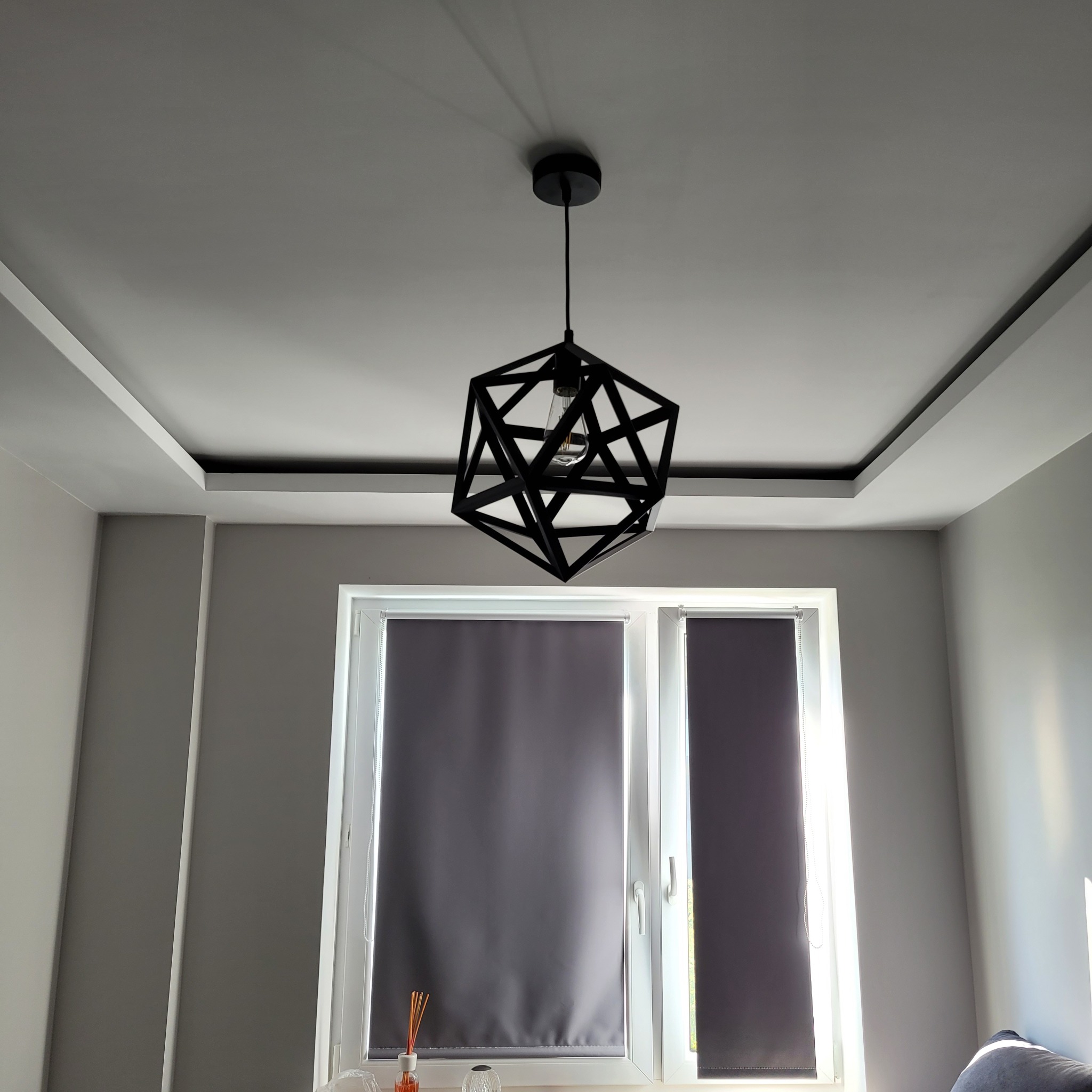 Czarny, geometryczny żyrandol wiszący na tle białego sufitu z dekoracyjną listwą i szarych ścian z oknem przysłoniętym roletami.