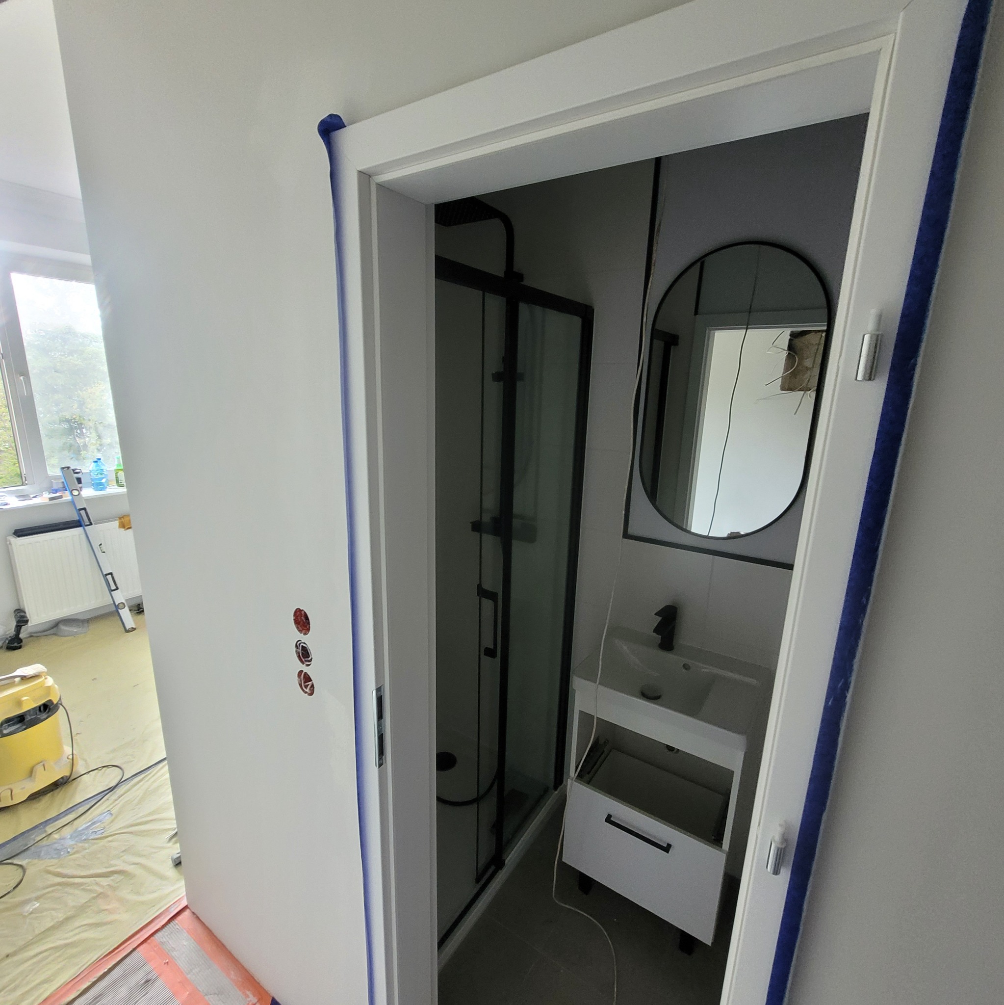 Wnętrze łazienki w trakcie remontu, widoczna kabina prysznicowa z czarnymi profilami, biała umywalka z szafką, lustro owalne w czarnej ramie, ściany przygotowane do malowania, zabezpieczone taśmą...