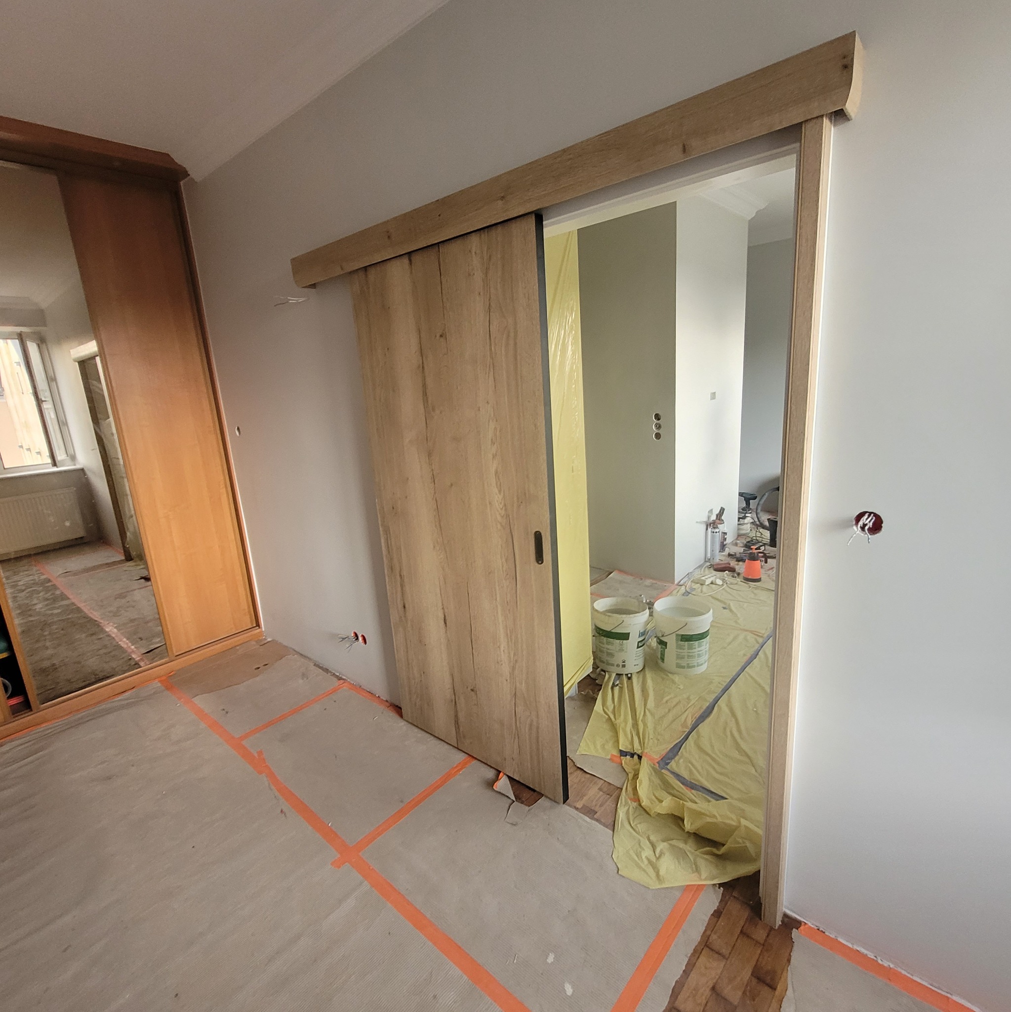 Montaż drewnianych drzwi przesuwnych w trakcie remontu, widoczne zabezpieczone podłogi i ściany, puszki elektryczne oraz wiaderka z farbą.