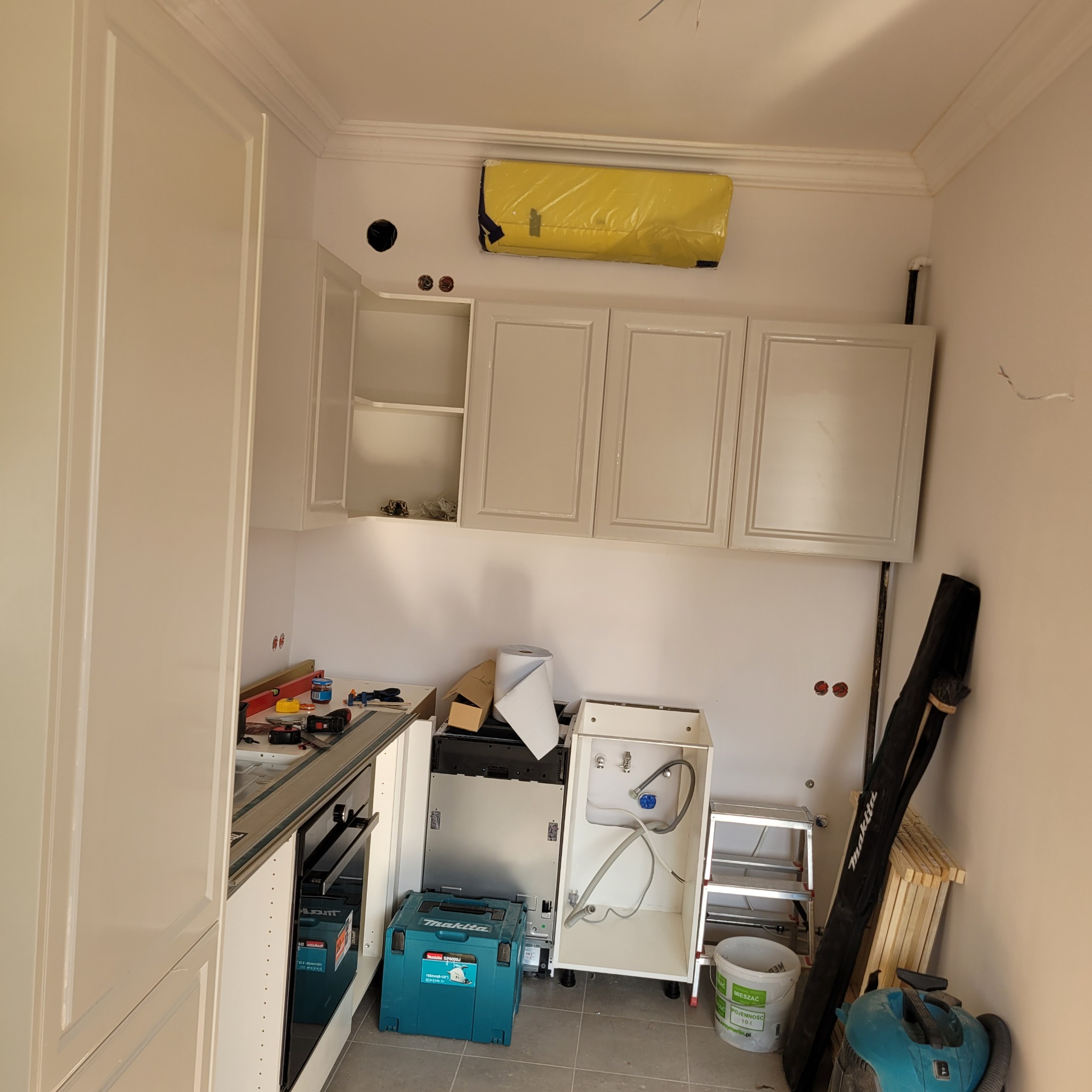 Wnętrze kuchni w trakcie montażu białych szafek wiszących i dolnych, widoczne narzędzia, niepodłączony zlew i osłonięty klimatyzator.