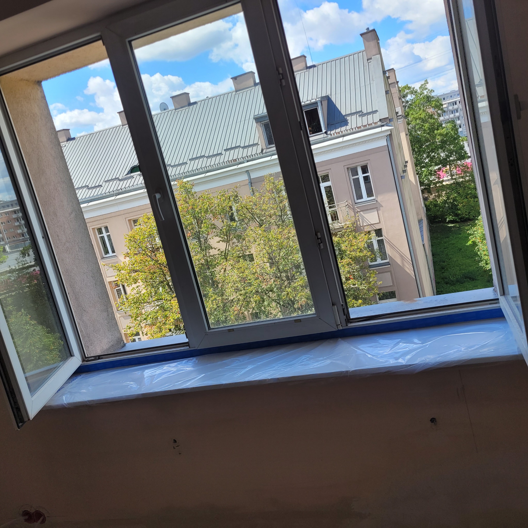 Okna PCV z widokiem na budynek i zieleń, zabezpieczony parapet folią malarską przed pracami wykończeniowymi.