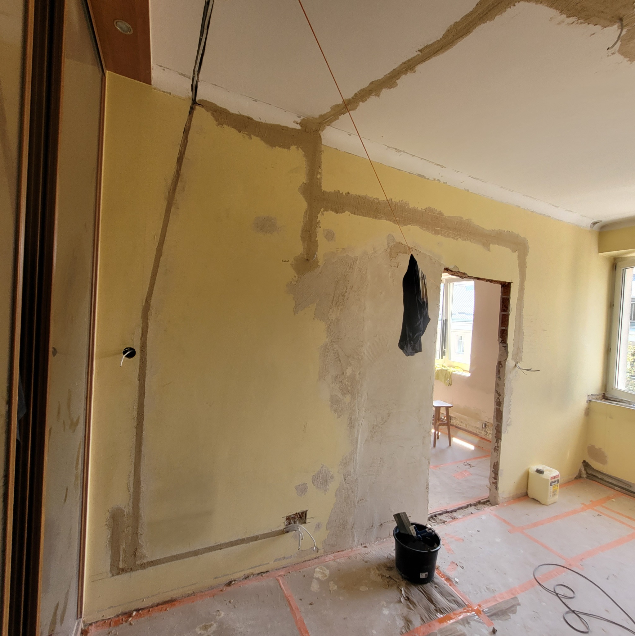 Wnętrze pokoju w trakcie remontu: ściany częściowo pokryte gładzią, widoczne instalacje elektryczne, podłoga zabezpieczona folią, wiadro z narzędziami w rogu.