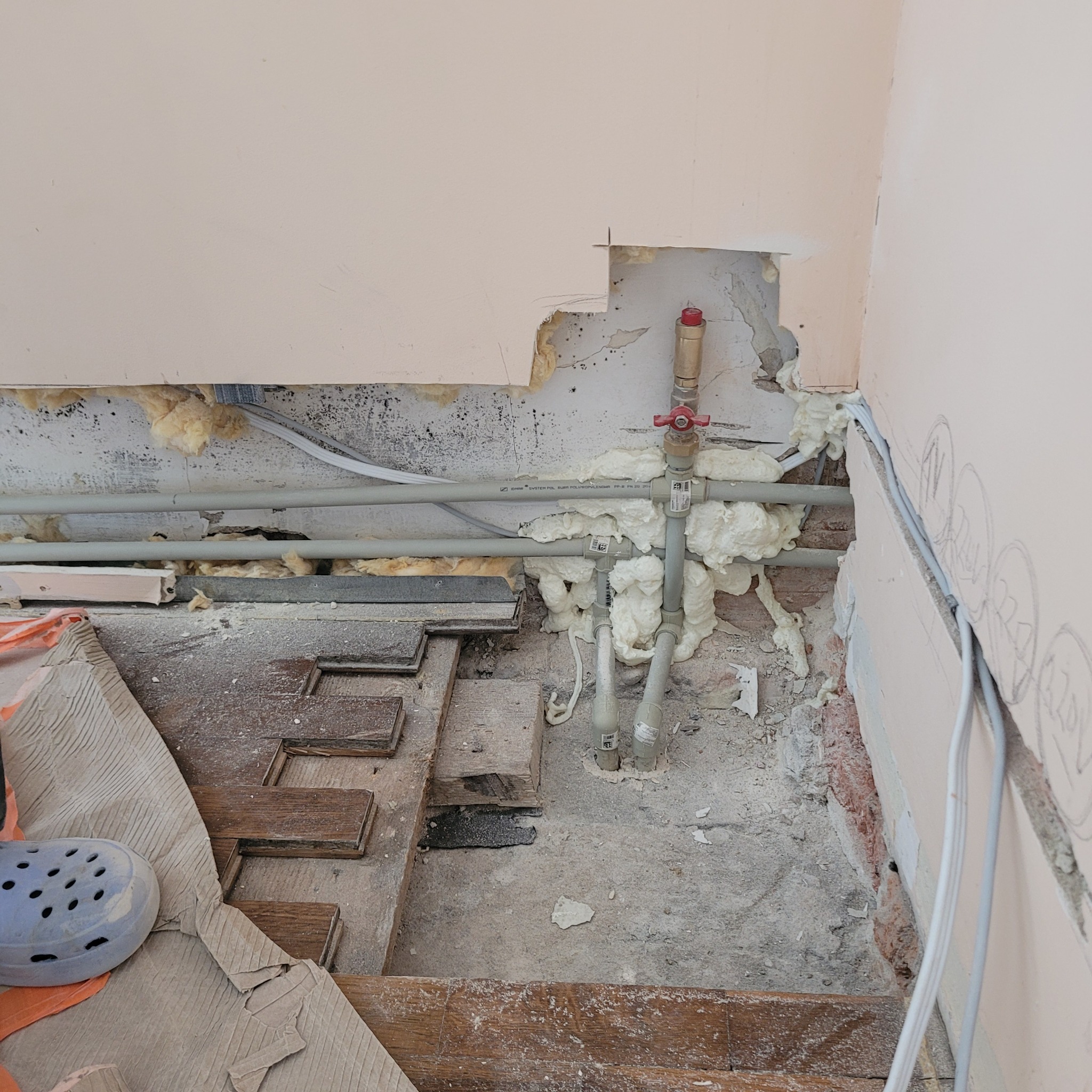 Fragment podłogi w trakcie demontażu starych paneli, widoczne rury instalacji wodnej zabezpieczone pianką montażową, odsłonięte kable elektryczne i fragment ściany z ubytkami tynku.