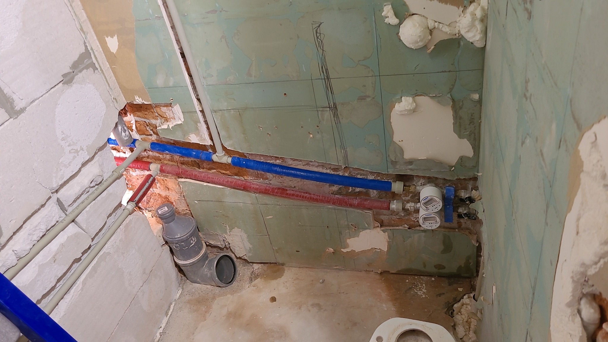 Łazienka w trakcie remontu: widoczne instalacje wodne (rury czerwone i niebieskie) oraz kanalizacyjne, ściany częściowo skute, z widocznymi warstwami farby i zaprawy, oraz pianka montażowa.