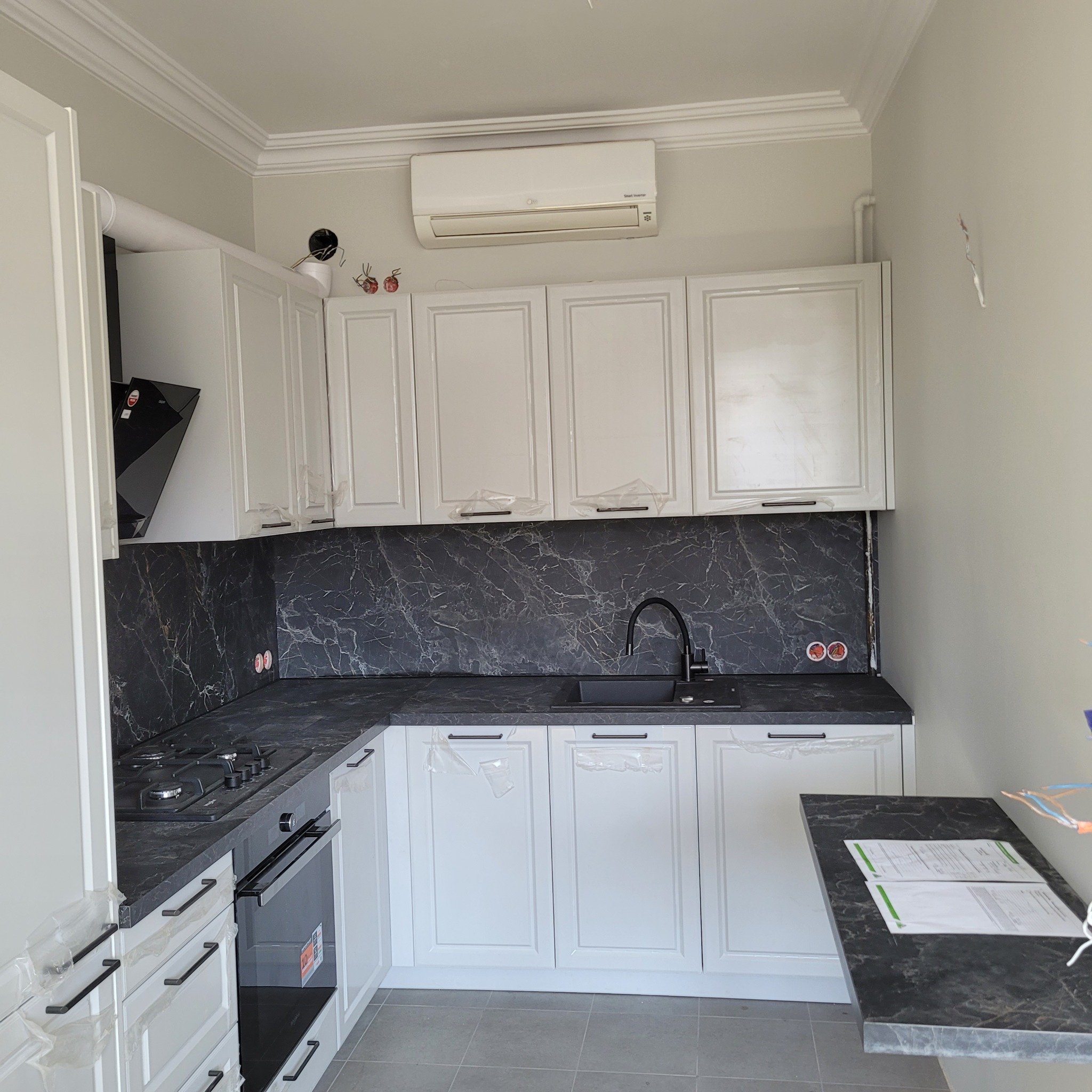 Nowoczesna kuchnia z białymi szafkami, czarnym blatem imitującym marmur i czarną armaturą, jeszcze w folii ochronnej, klimatyzator na ścianie.