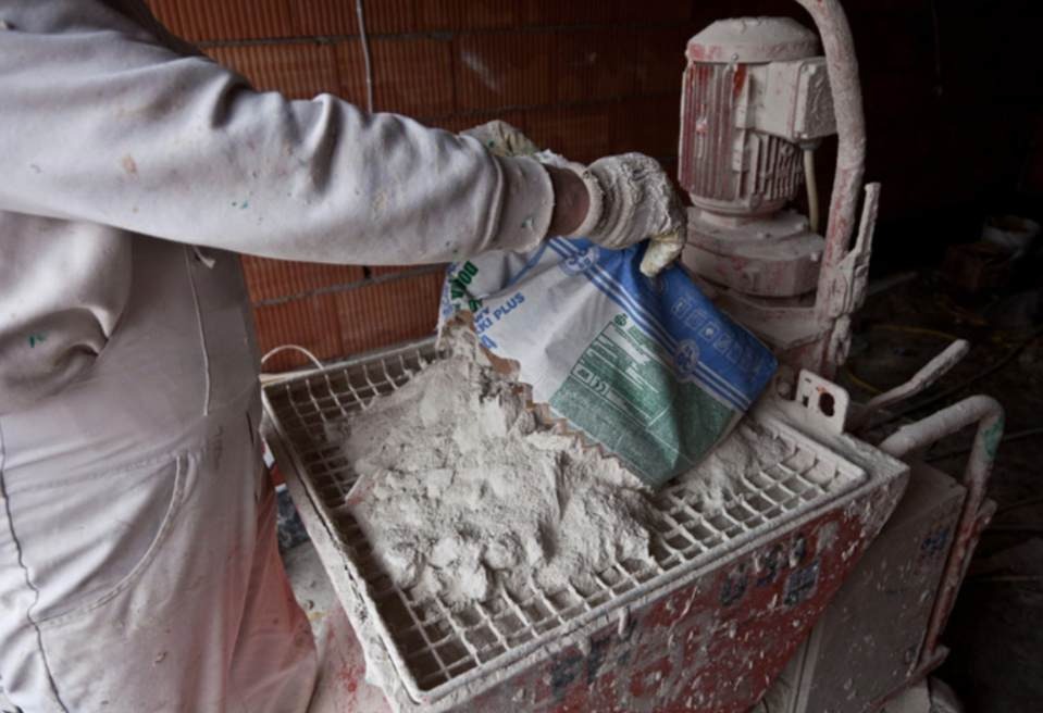 Pracownik w Łyse dosypuje suchą mieszankę z worka do brudnej maszyny do tynkowania, widoczne cegły w tle.