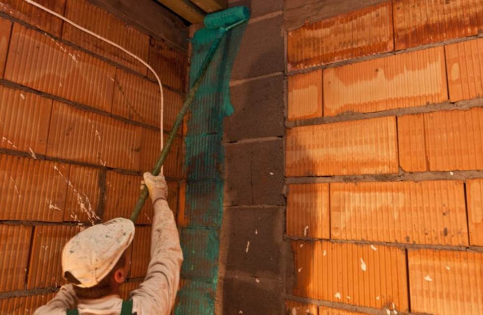 Mężczyzna w roboczym ubraniu maluje narożnik ściany z cegieł ceramicznych wałkiem na długim uchwycie, widoczne ślady zaprawy i instalacja elektryczna.