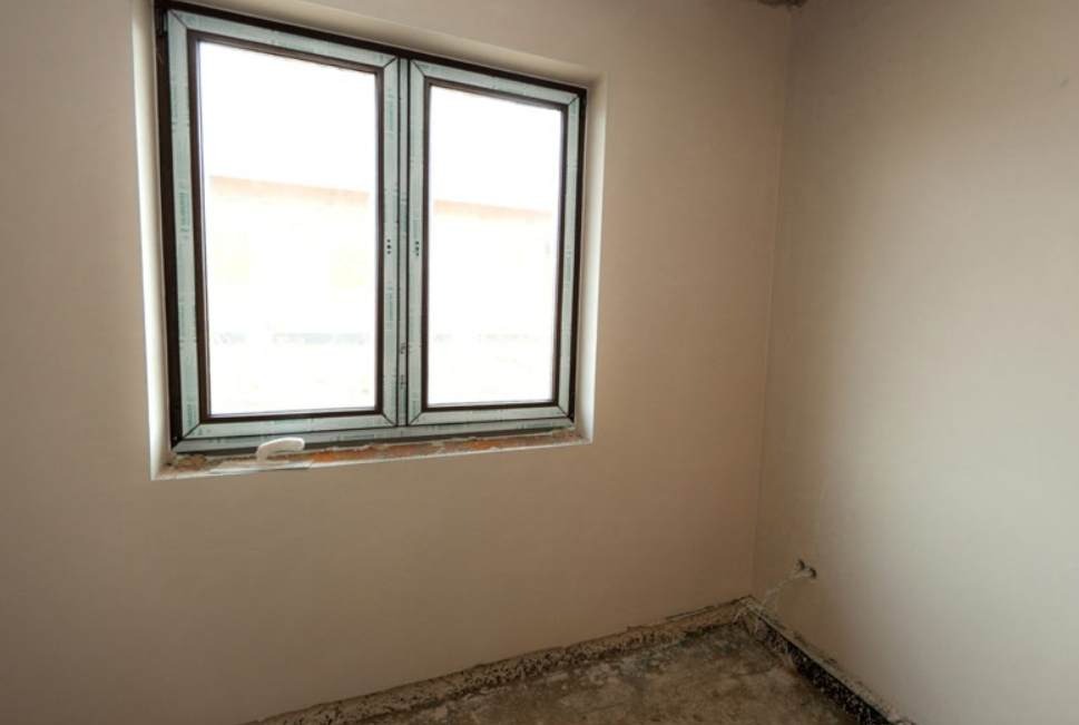 Wnętrze pokoju po gładziowaniu ścian i montażu okna PCV z brązową ramą; widoczny fragment podłogi przed położeniem posadzki.