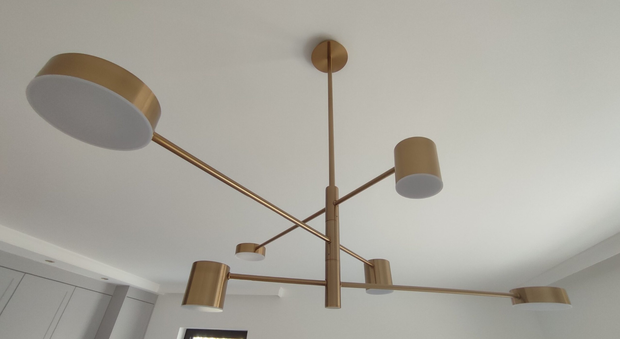 Nowoczesna, złota lampa sufitowa z pięcioma punktami świetlnymi o różnej wielkości, zamontowana na białym suficie w pomieszczeniu z szarymi szafkami w tle.