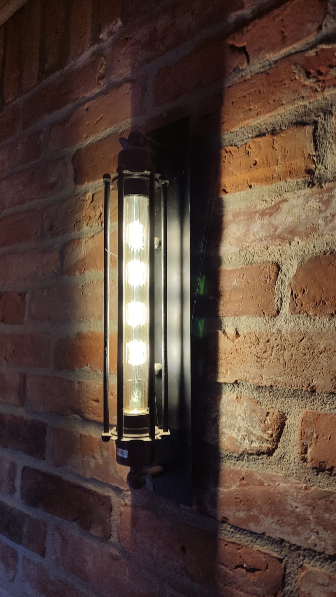 Kinkiet w stylu industrialnym z podłużną żarówką z czterema punktami świetlnymi, zamontowany na ścianie z czerwonej cegły.