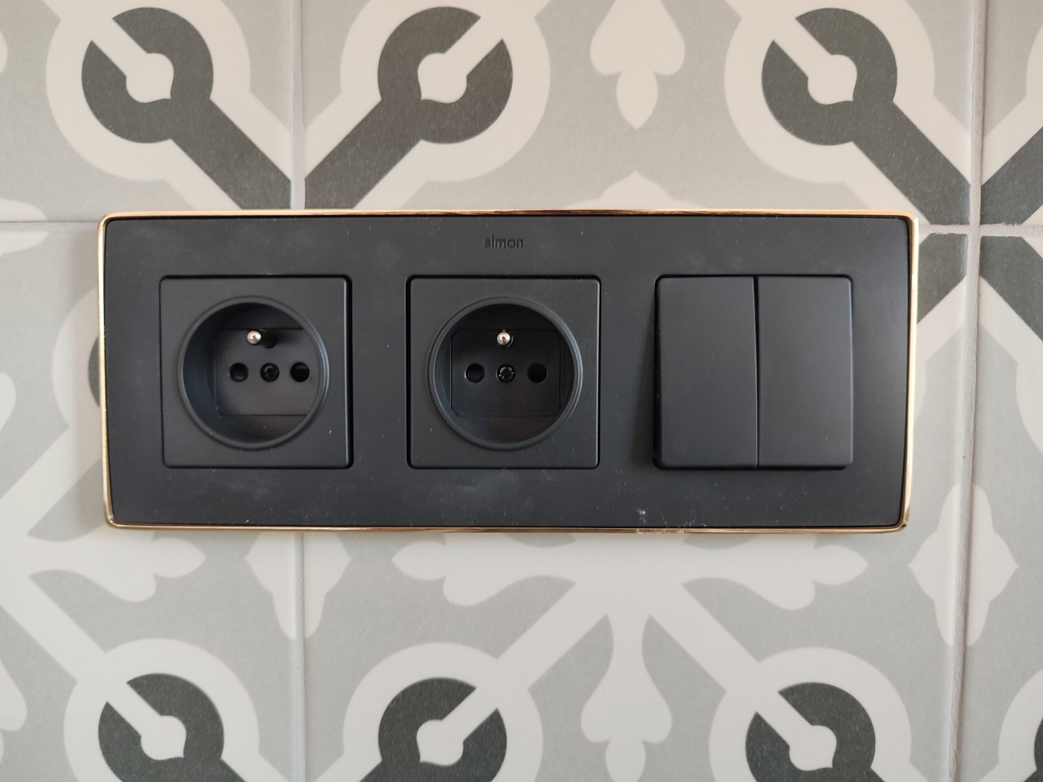 Czarny podwójny kontakt i włącznik światła marki Simon z cienką złotą ramką, zamontowane na ścianie z szaro-białymi wzorzystymi płytkami.
