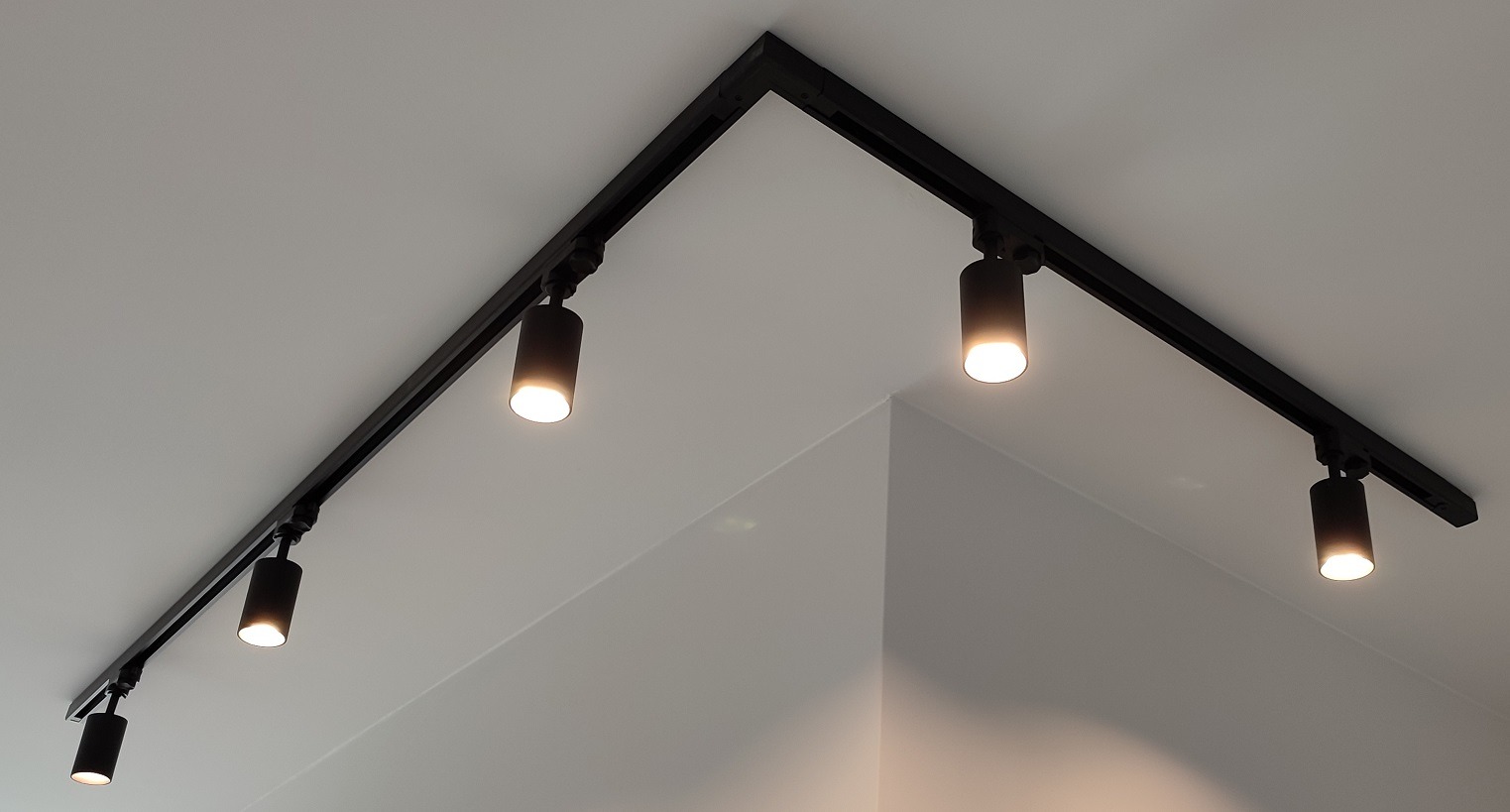 Czarna szyna oświetleniowa z czterema reflektorami cylindrycznymi zamontowana na białym suficie w rogu pomieszczenia.