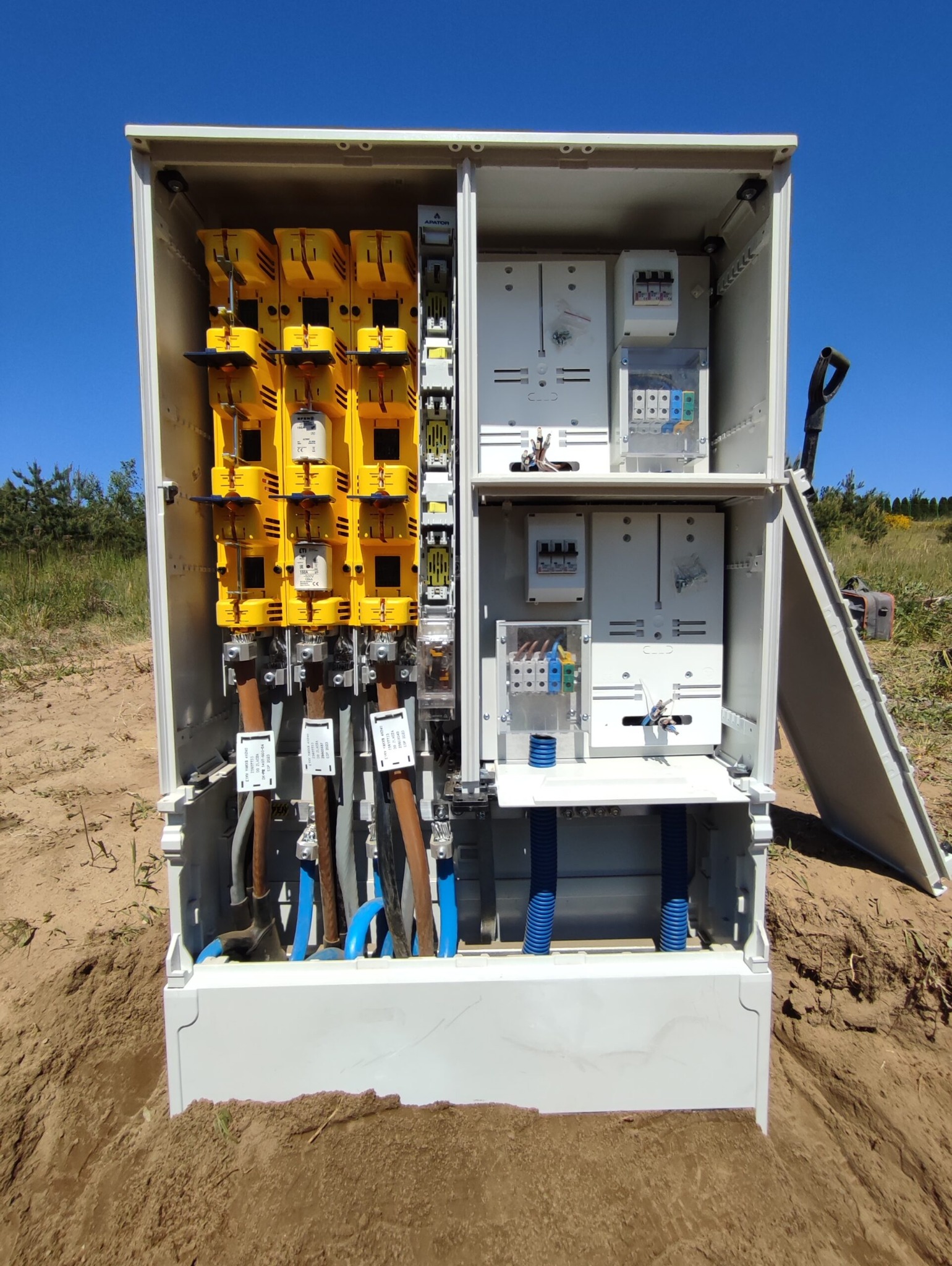 Otwarta szafa elektryczna z żółtymi bezpiecznikami i niebieskimi rurami kablowymi, umieszczona na ziemi w plenerze.