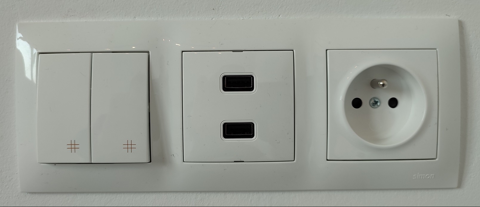 Biały panel ścienny z dwoma włącznikami światła z symbolem kratki, podwójnym portem USB i gniazdkiem elektrycznym typu europejskiego, marki Simon.