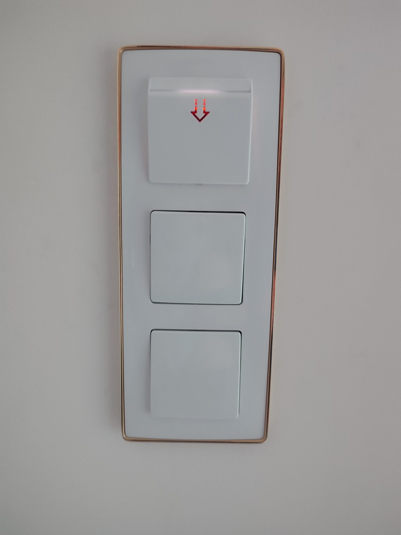 Biały panel z trzema włącznikami, górny z podświetlaną czerwoną strzałką w dół, otoczony złotą ramką, zamontowany na jasnej ścianie.