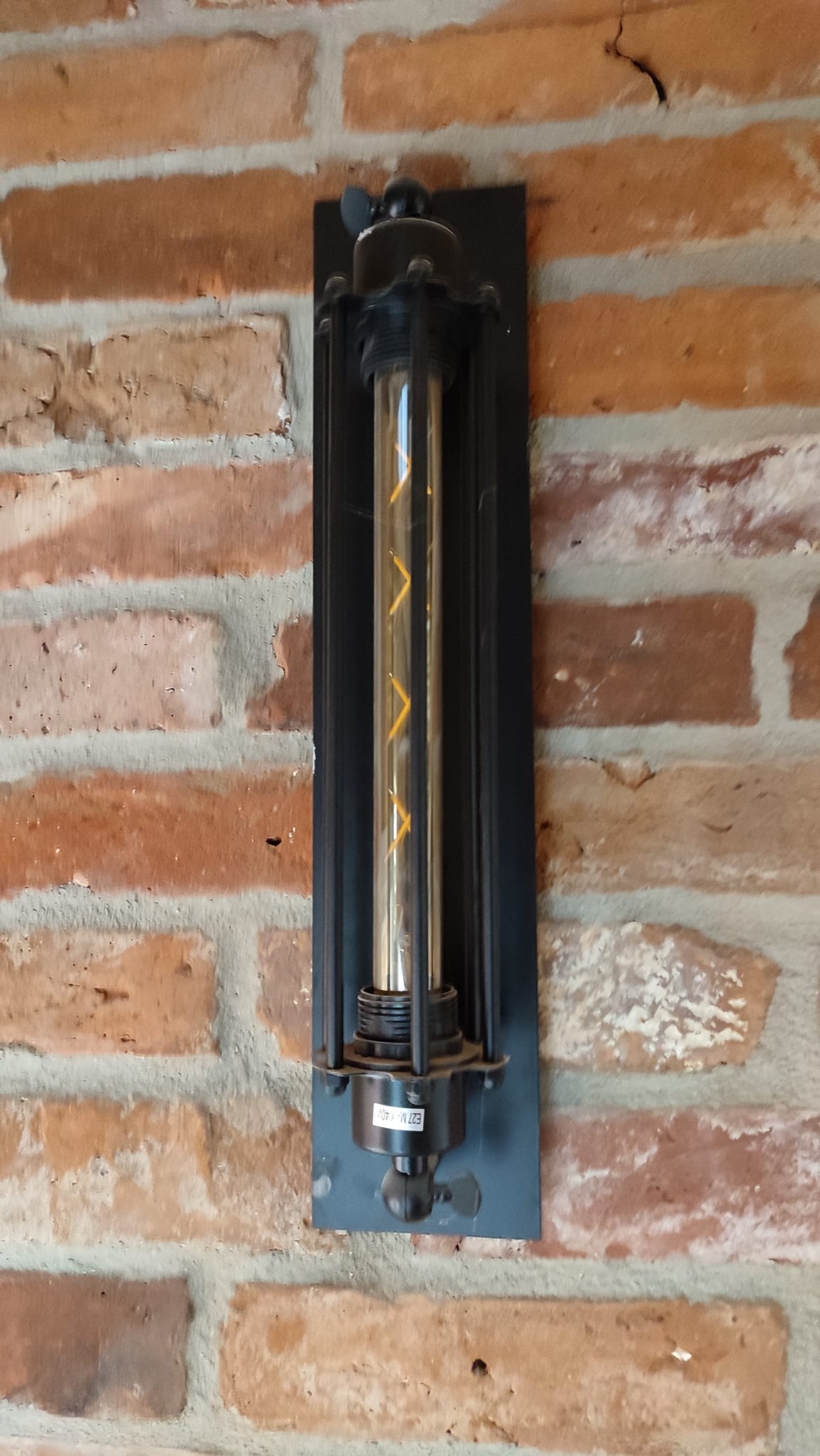 Czarny, metalowy kinkiet z żarówką filamentową o spiralnym wzorze, zamontowany na ścianie z czerwonej cegły w Słupsku. Widoczne detale mocowania i specyfikacja żarówki.