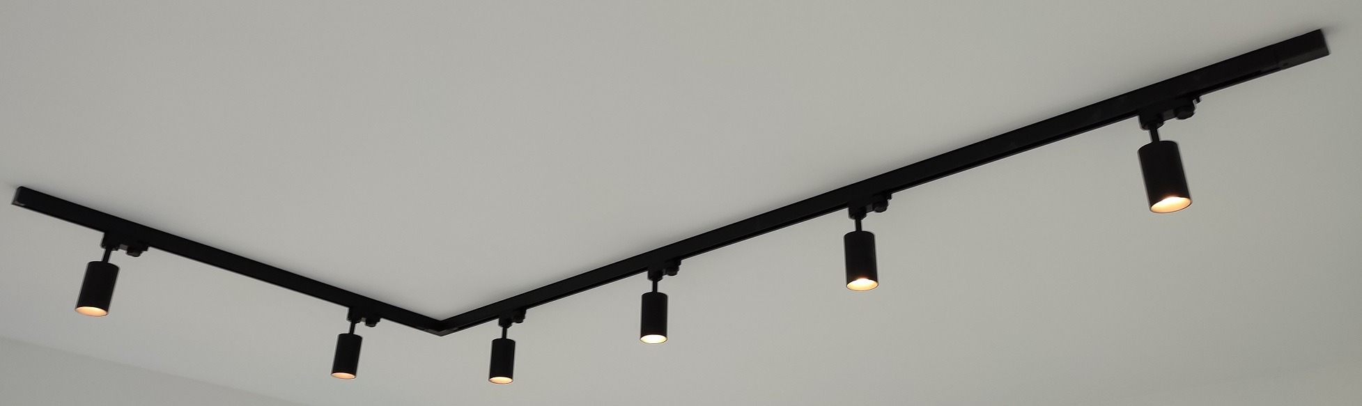 Czarna, szynowa instalacja oświetleniowa z reflektorami cylindrycznymi zamontowana na białym suficie, ułożona w kształt litery V.