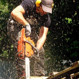 Black Forest - Mężczyzna w ochronnikach słuchu tnący drewno piłą łańcuchową, w powietrzu unoszą się wióry, w tle zieleń.