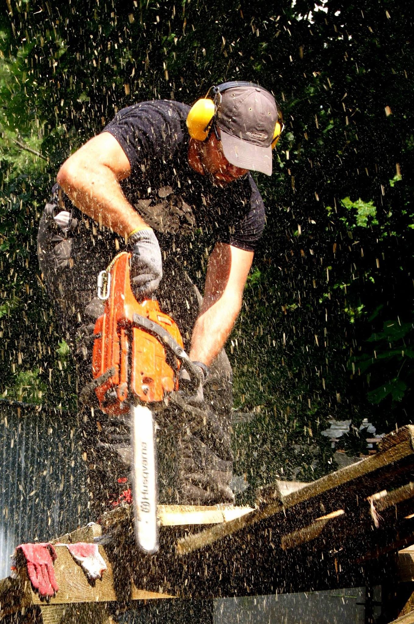 Mężczyzna w ochronnikach słuchu tnący drewno piłą łańcuchową, w powietrzu unoszą się wióry, w tle zieleń.