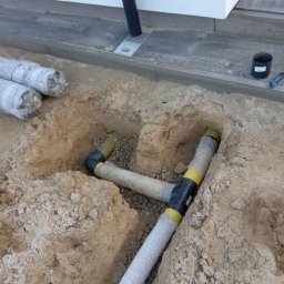 KIKI USŁUGI MINI KOPARKĄ - Układanie rur drenażowych w wykopie obok budynku, z widocznymi elementami łączeniowymi i obsypką żwirową. Na powierzchni ziemi leżą rolki materiału izolacyjnego.