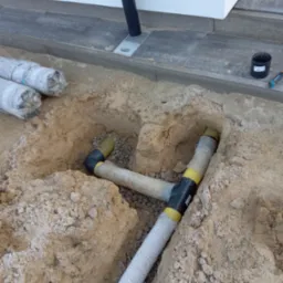 Układanie rur drenażowych w wykopie obok budynku, z widocznymi elementami łączeniowymi i obsypką żwirową. Na powierzchni ziemi leżą rolki materiału izolacyjnego.