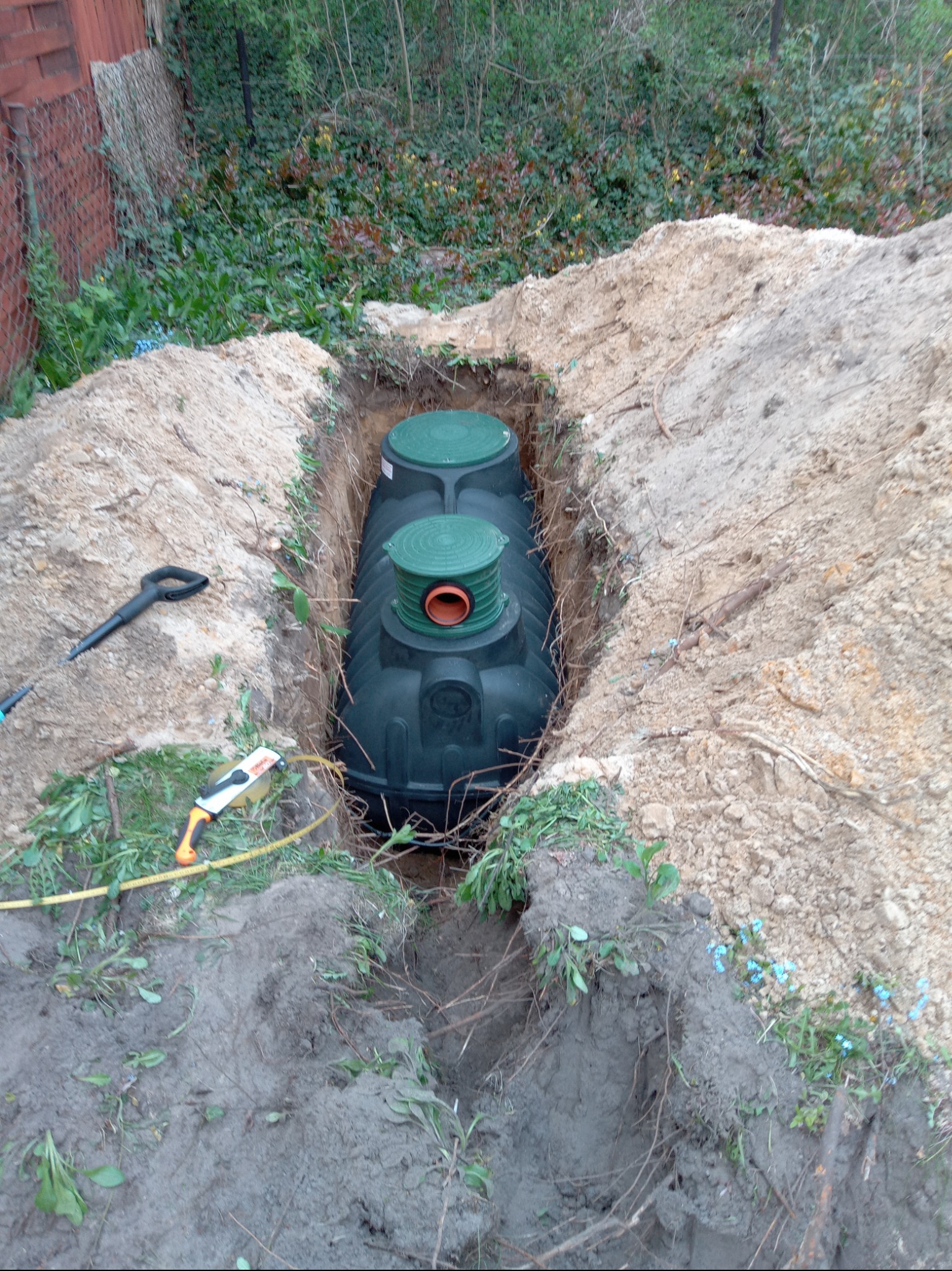 Zielony, plastikowy zbiornik na szambo umieszczony w wykopie ziemnym, z widocznymi narzędziami: łopatą i miarką zwijaną. Tło stanowi zielona roślinność.
