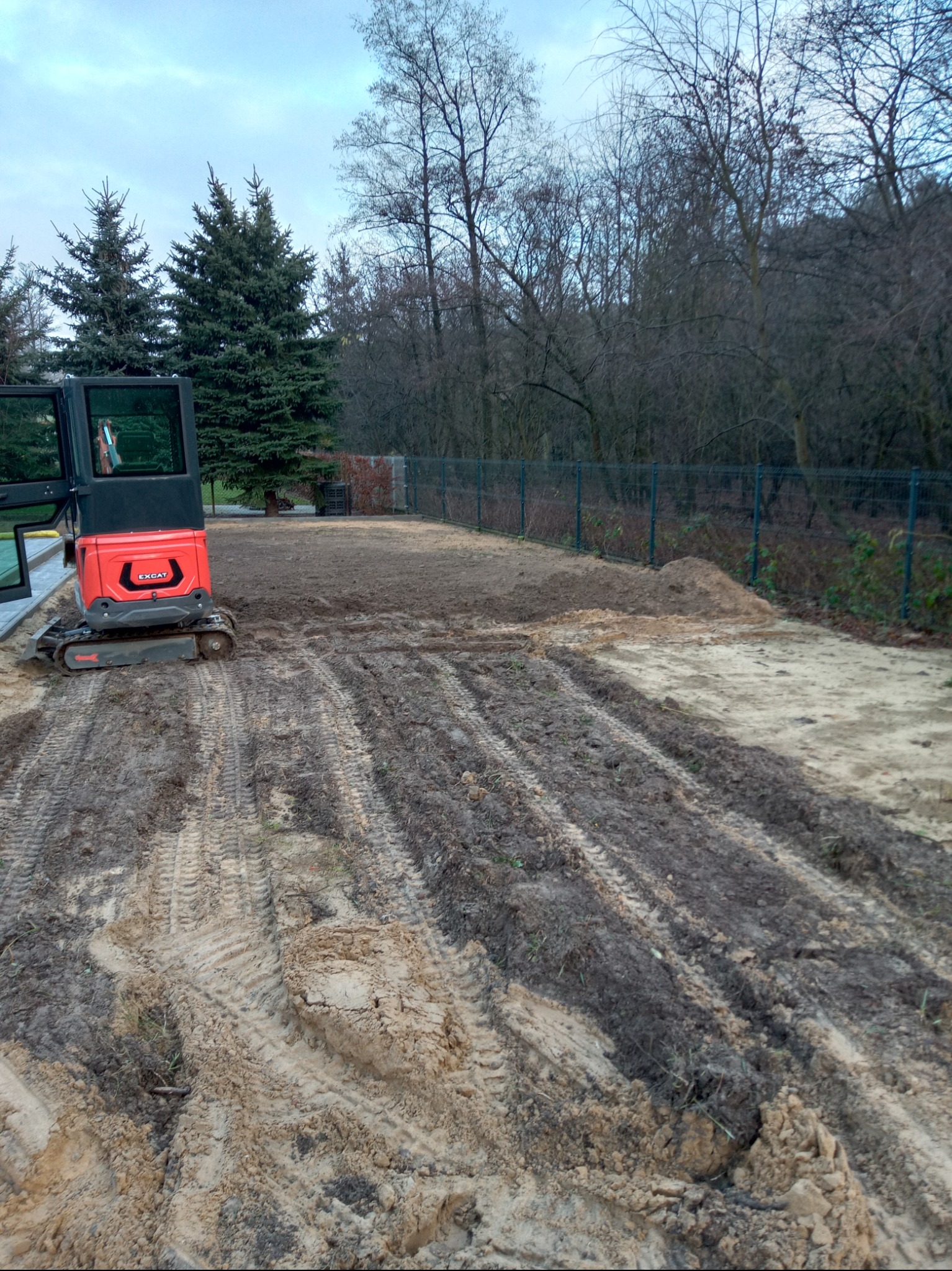 Czerwona mini koparka EXCAT na placu budowy z wykopanymi rowami w ziemi, obok zielone drzewa i siatka ogrodzeniowa. Przygotowanie terenu pod budowę.