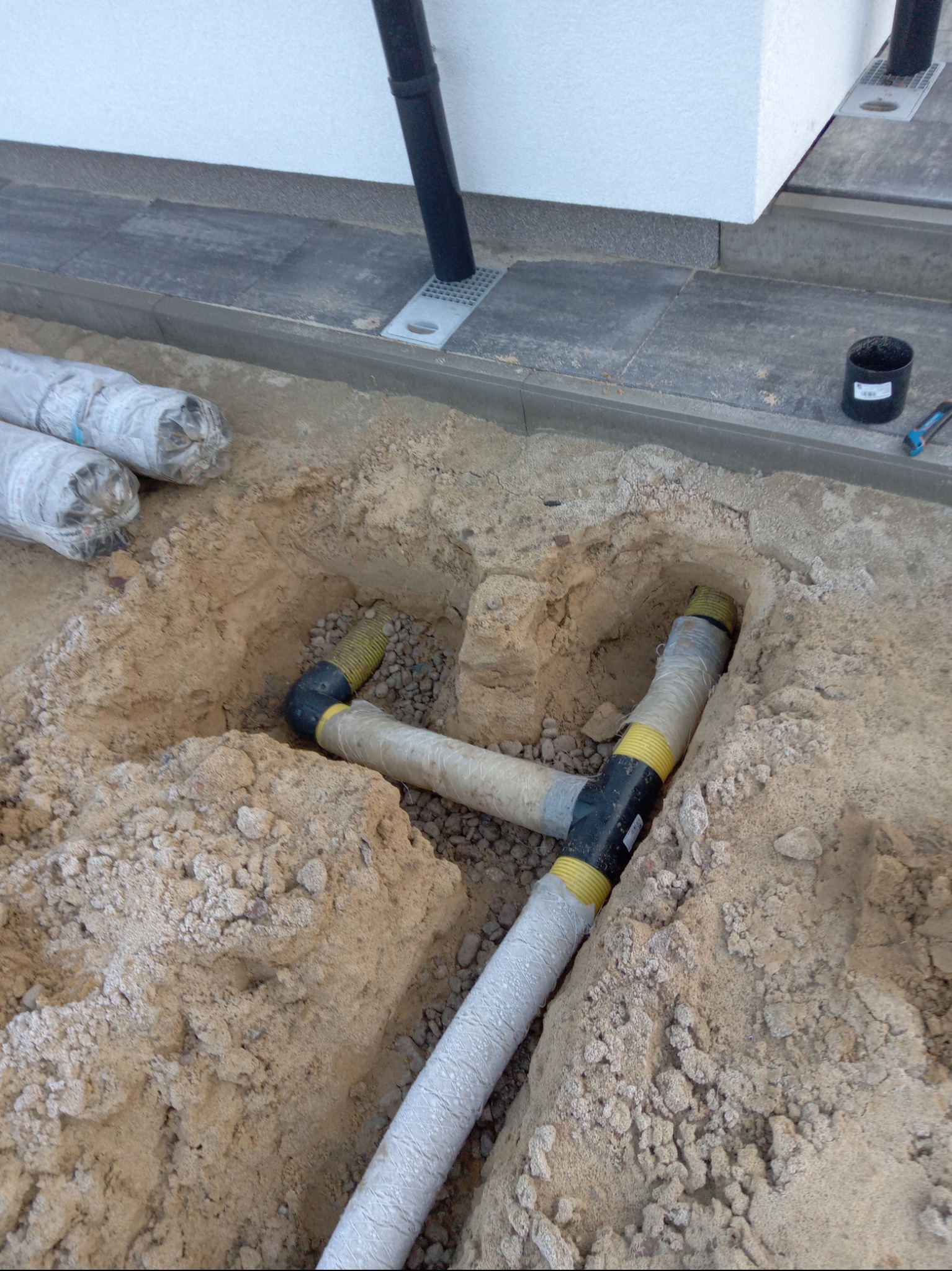 Układanie rur drenażowych w wykopie obok budynku, z widocznymi elementami łączeniowymi i obsypką żwirową. Na powierzchni ziemi leżą rolki materiału izolacyjnego.