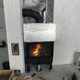 INHYDRO USŁUGI HYDRAULICZNE GRZEGORZ KUKUCZKA - Wnętrze w trakcie montażu kominka z otwartym paleniskiem, widoczne rury odprowadzające spaliny, obudowa w trakcie wykańczania, na podłodze kartony i narzędzia.