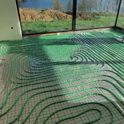 INHYDRO USŁUGI HYDRAULICZNE GRZEGORZ KUKUCZKA - Zielone rurki ogrzewania podłogowego ułożone na siatce w surowym wnętrzu z dużymi oknami, widok na zewnątrz na zieleń i jezioro, widoczny fragment ściany z otworem.