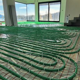 INHYDRO USŁUGI HYDRAULICZNE GRZEGORZ KUKUCZKA - Układanie zielonych rur ogrzewania podłogowego na folii z siatką w przestronnym, jasnym pomieszczeniu z dużymi oknami i widocznymi schodami.