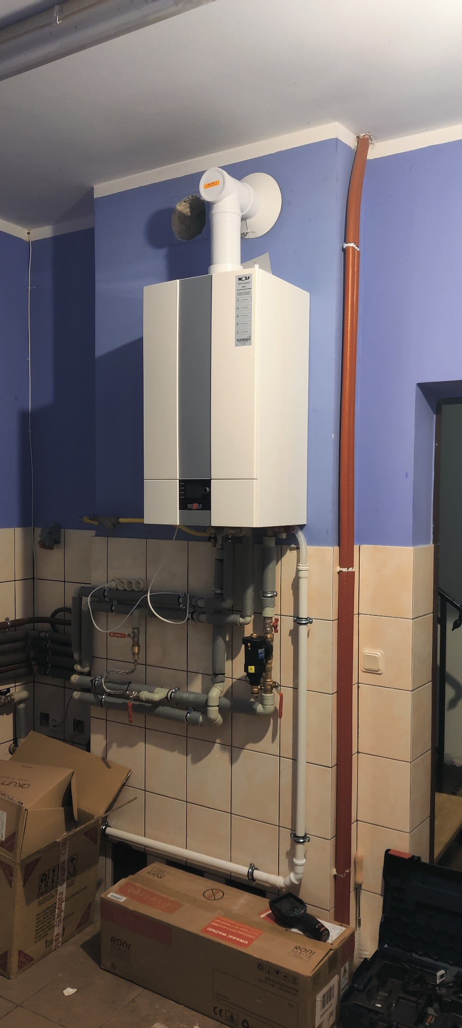 Nowy, biały piec gazowy marki Wolf zamontowany na ścianie z niebieską górną częścią i beżową dolną z kafelkami. Widoczne rury i elementy instalacji, kartony po urządzeniach na podłodze, narzędzia...