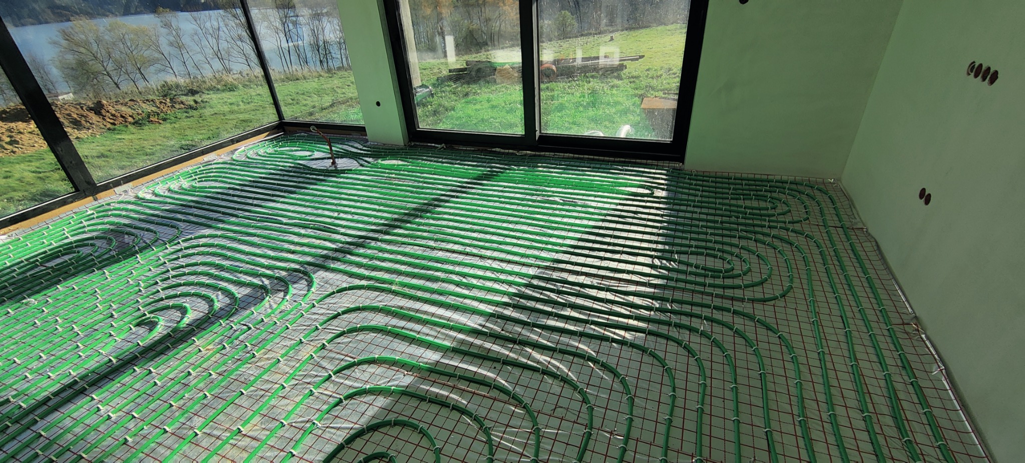 Zielone rury ogrzewania podłogowego ułożone na siatce w jasnym pomieszczeniu z dużymi oknami, widok na zewnątrz z jeziorem i zielenią.