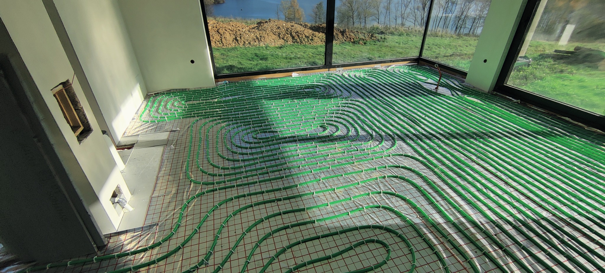 Zielone rurki ogrzewania podłogowego ułożone na siatce w surowym wnętrzu z dużymi oknami, widok na zewnątrz na zieleń i jezioro, widoczny fragment ściany z otworem.