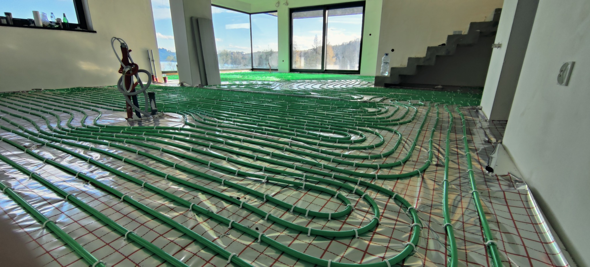 Układanie zielonych rur ogrzewania podłogowego na folii z siatką w przestronnym, jasnym pomieszczeniu z dużymi oknami i widocznymi schodami.